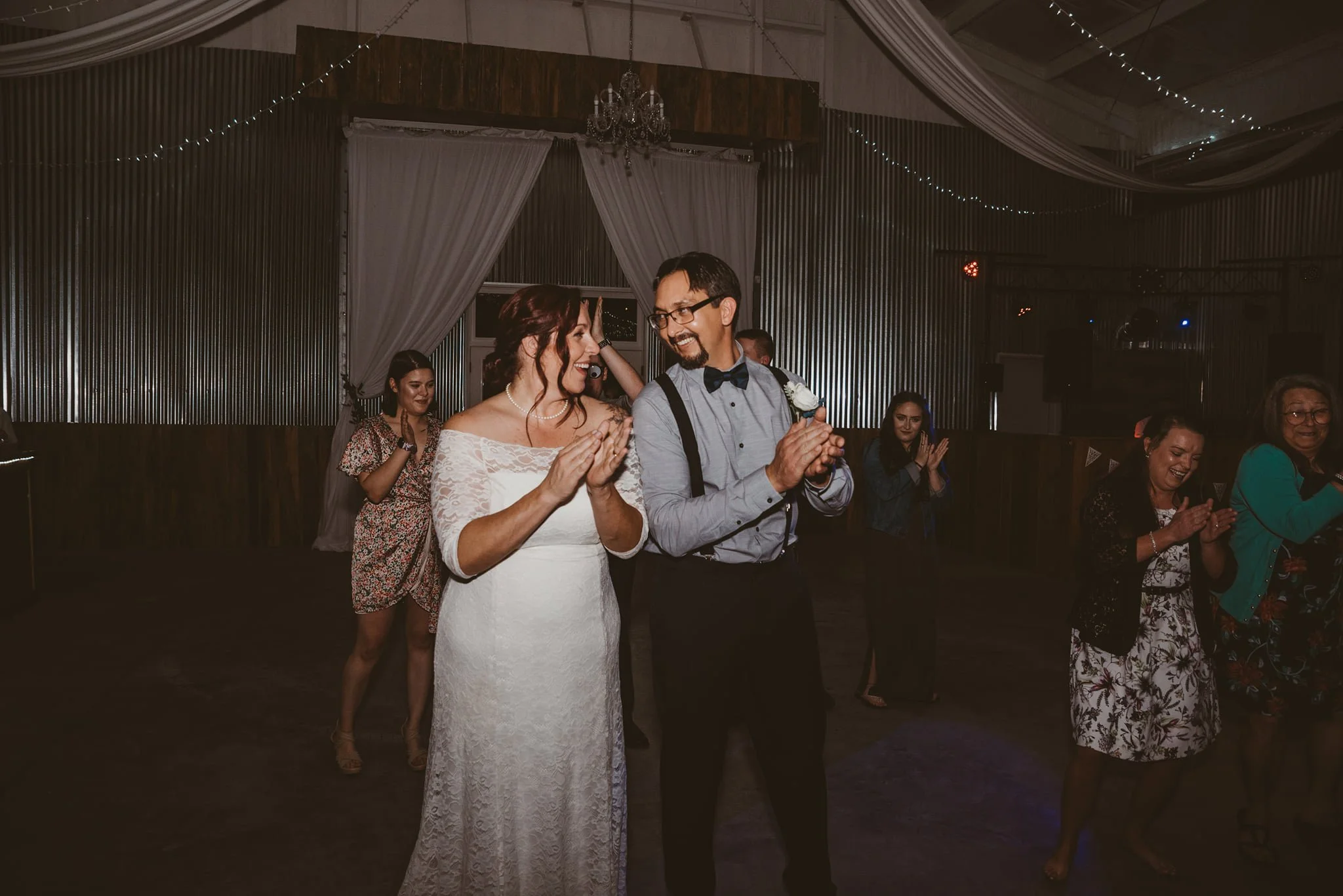 Bride and groom dancing at their wedding reception with guests clapping and smiling around them in rustic decorated hall.