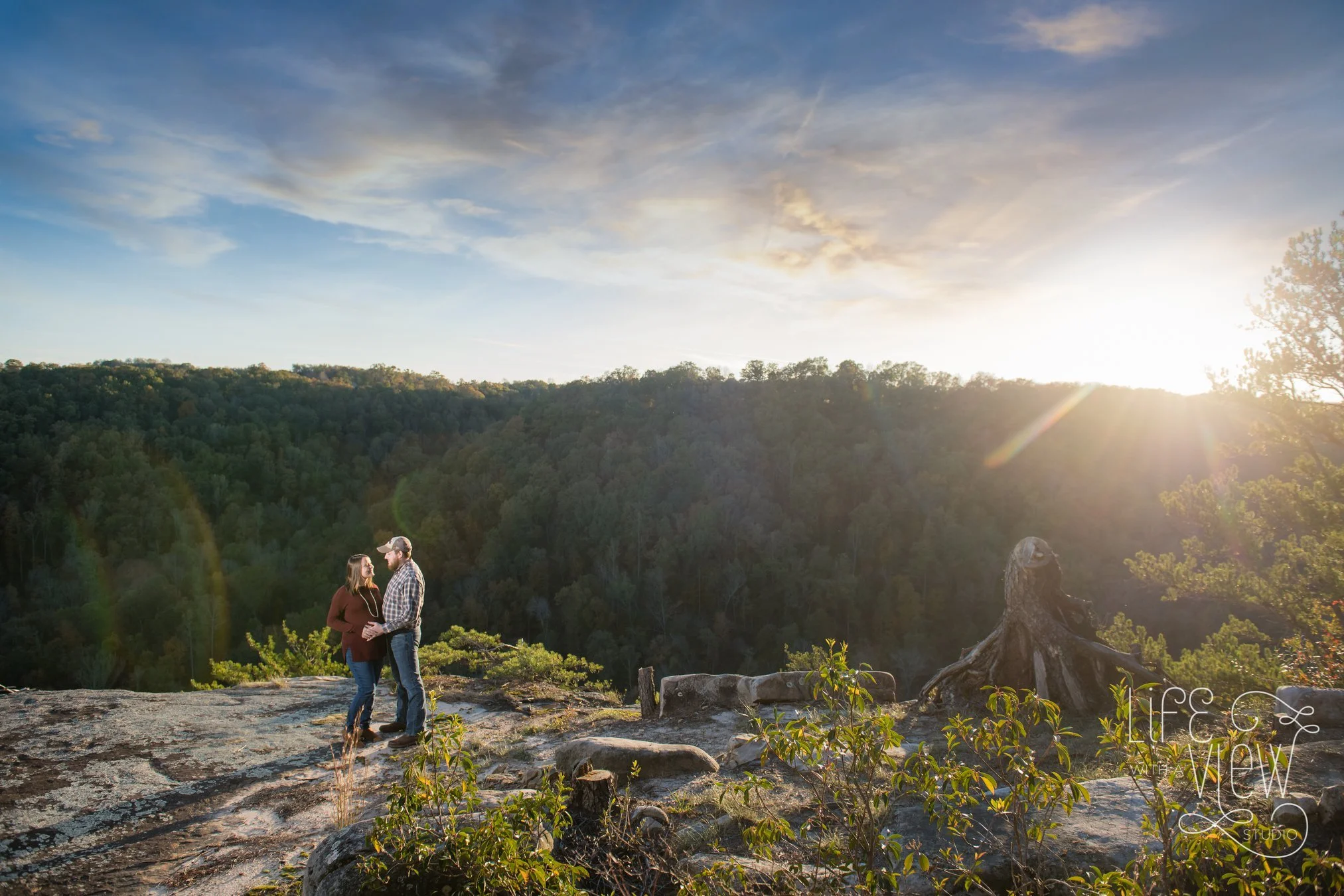 A couple standing on a rocky overlook at sunset with a view of a forested valley.