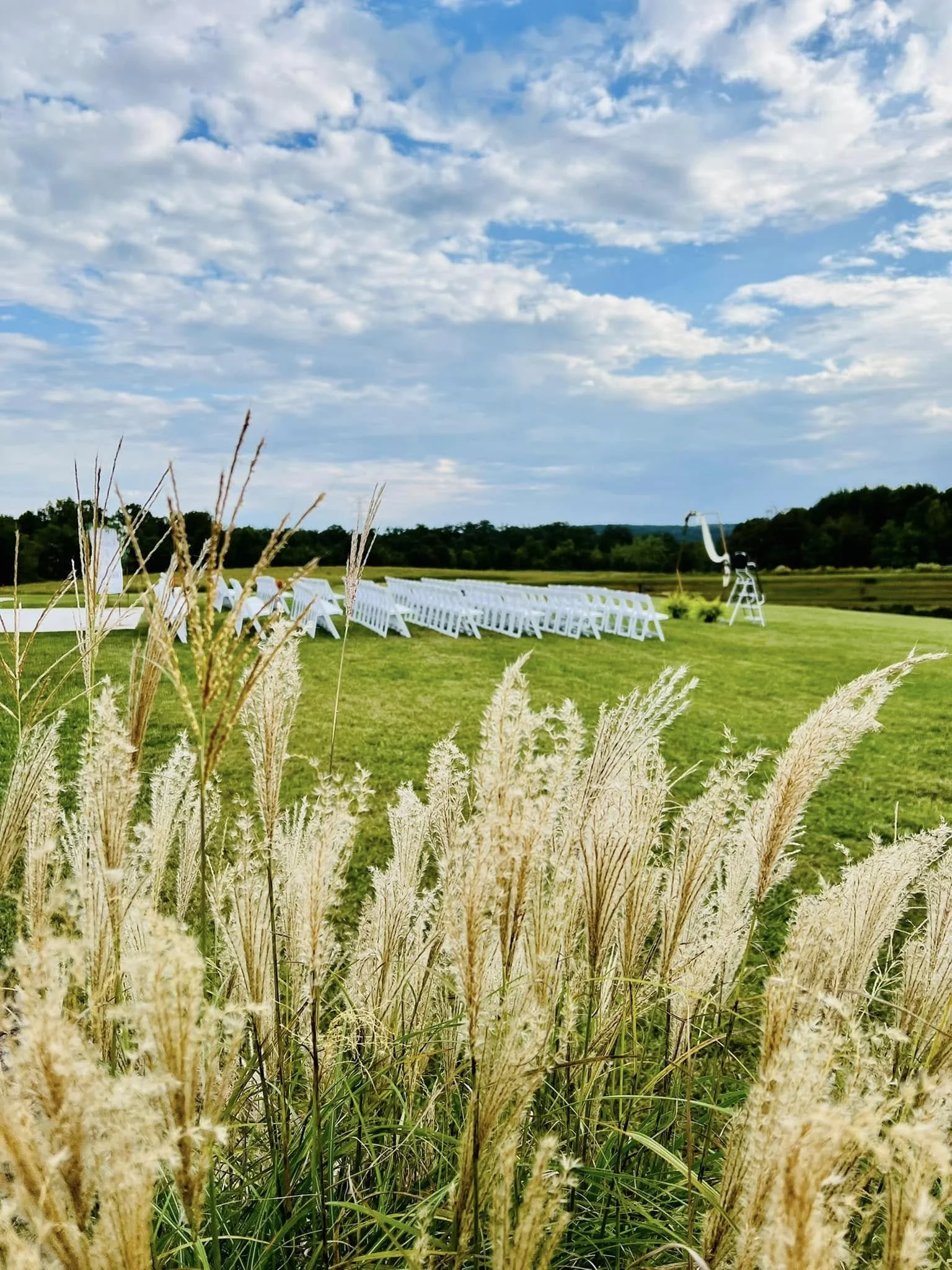 Outdoor wedding setup on a grassy field with white chairs facing a telescope under a partly cloudy sky.