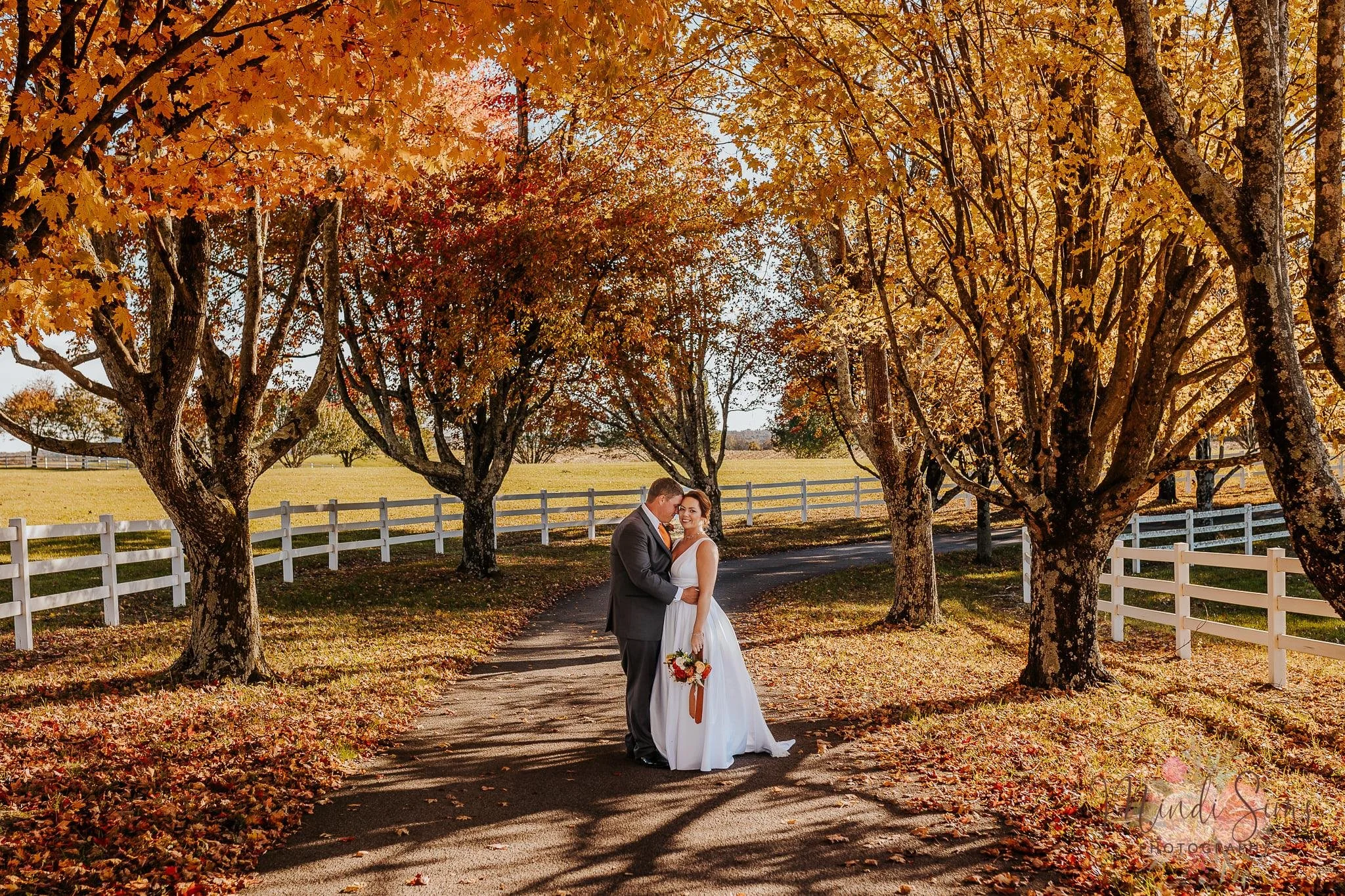 A wedding couple standing on a dirt path surrounded by trees with autumn leaves, white fencing, and a grassy field in the background, during daytime.