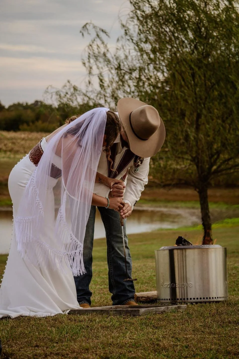 A couple dressed in wedding attire engaging in a fire ceremony outdoors, with trees and a pond in the background.