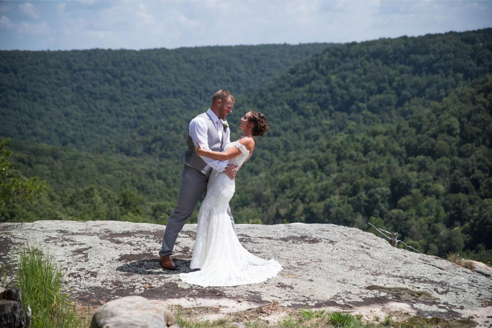 A bride and groom on a rocky outdoor area with a forested mountain background, embracing and smiling at each other, during their wedding photoshoot.