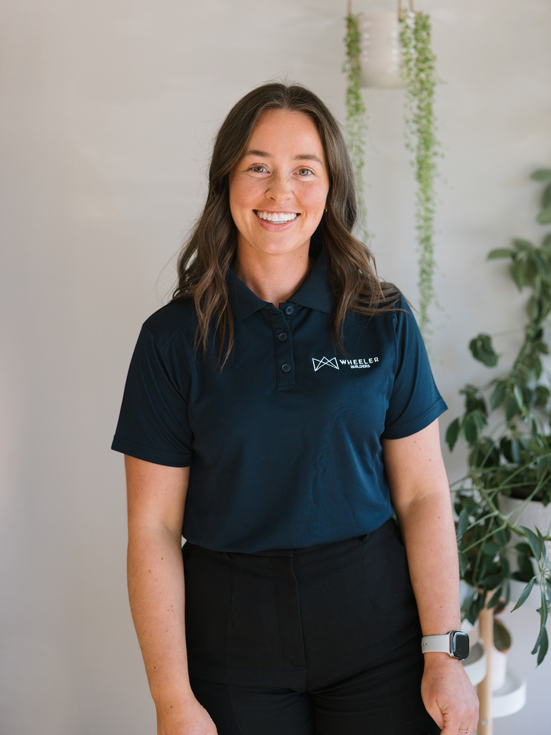 A woman in a navy blue polo shirt standing indoors next to a plant.