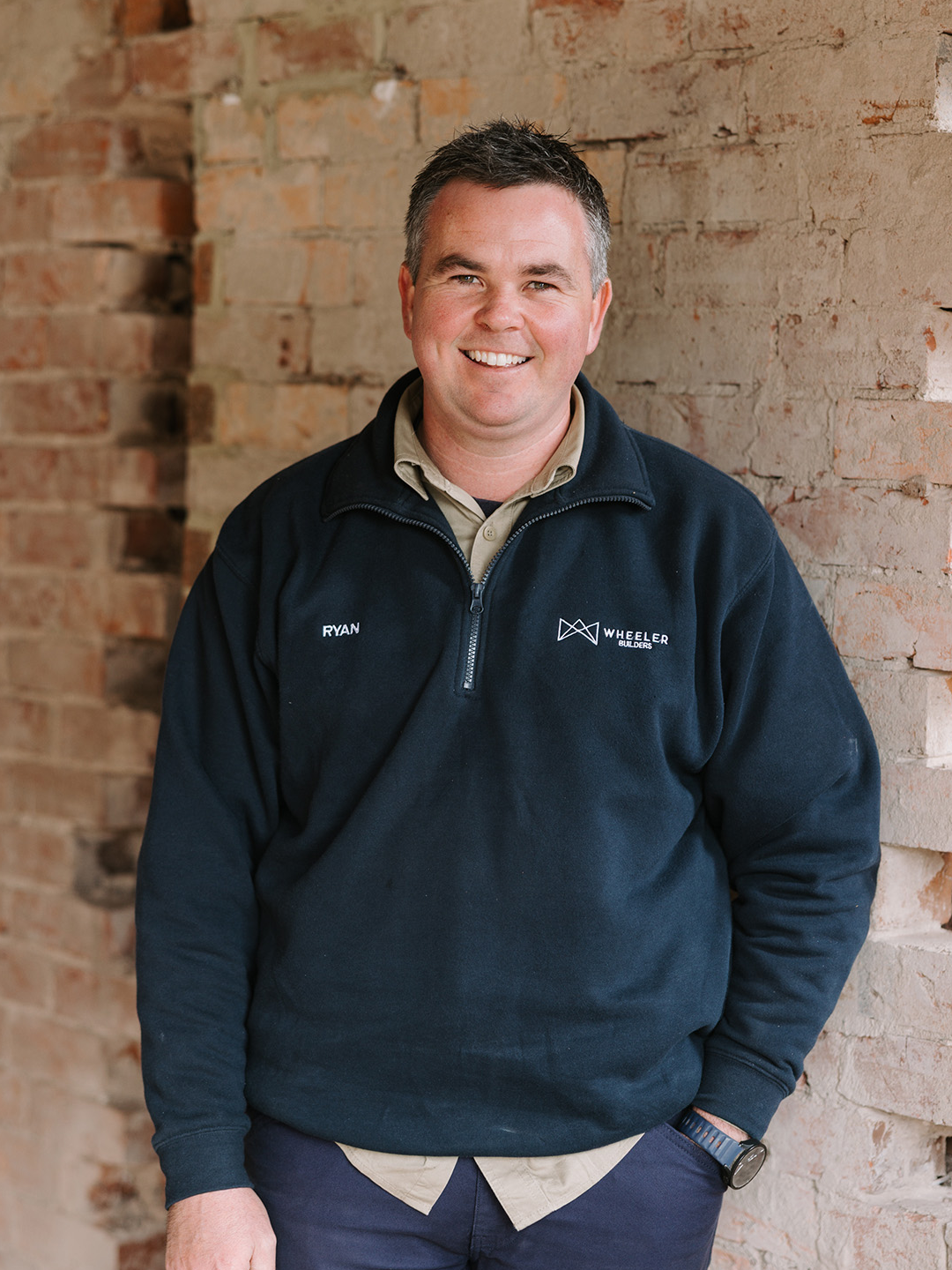 A man smiling, wearing a navy fleece with the name 'Ryan' and 'Wheeler Builders' embroidered on it, standing against a brick wall.
