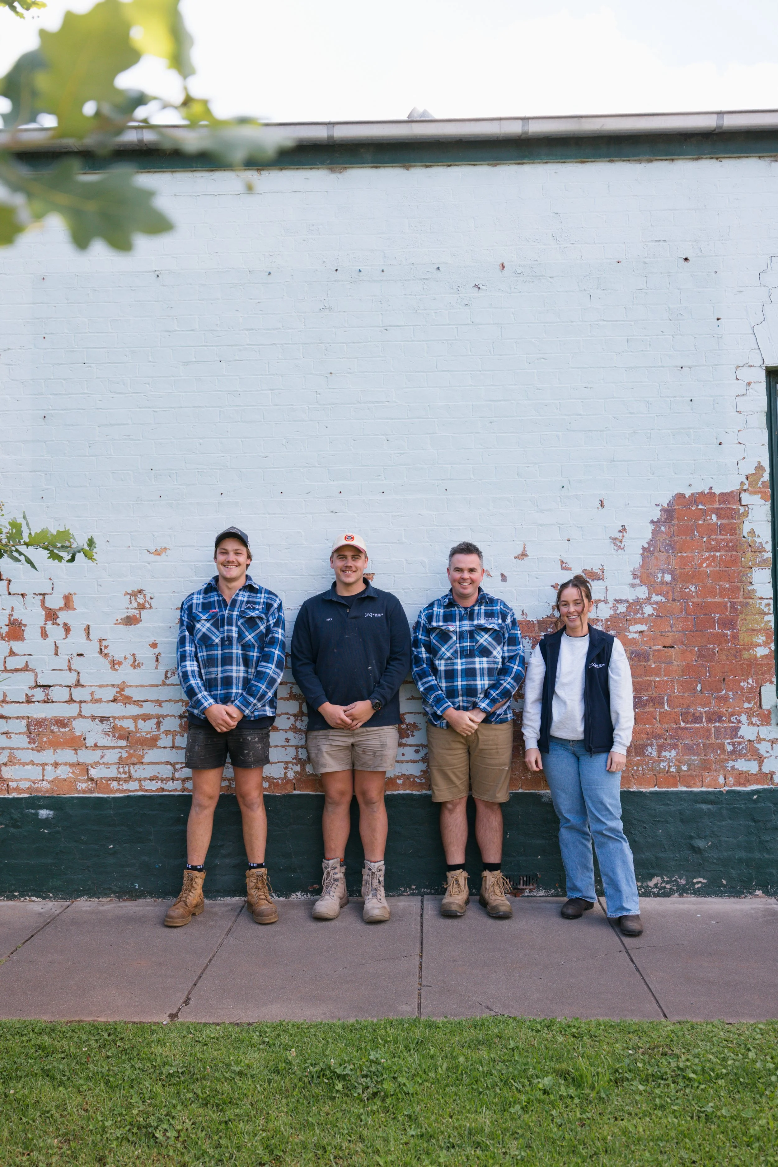 Four young adults standing in front of a brick wall with peeling paint, smiling at the camera. They are dressed casually, with three wearing plaid shirts and boots, and one wearing a jacket and jeans. One is wearing a cap and another has a hoodie.