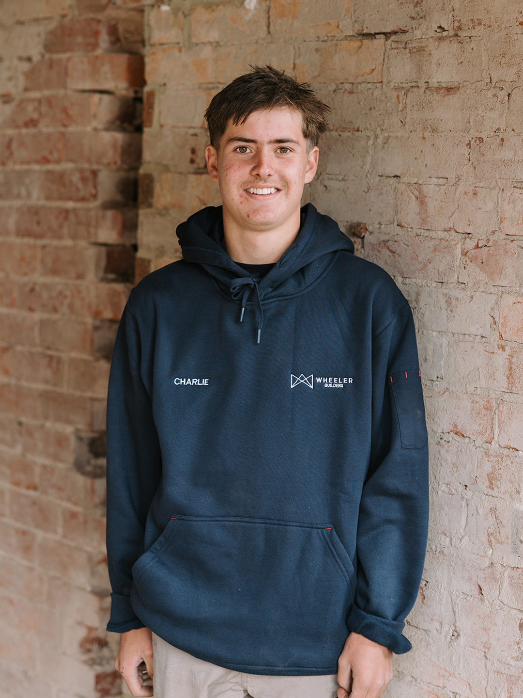 A young man with short brown hair and a slight smile, wearing a dark blue hoodie with embroidered name 'Charlie' and logo 'Wheeler Builders', standing against a brick wall.