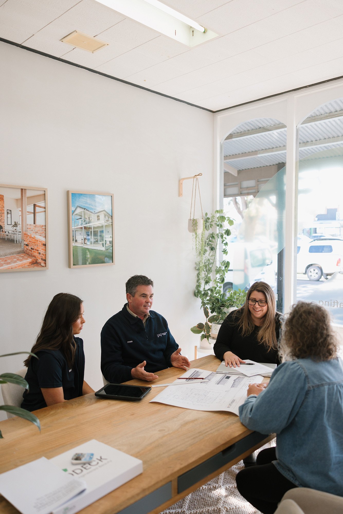 A group of people sitting around a wooden table, engaged in conversation, with documents and a tablet on the table inside a bright room with large windows, white walls, and framed pictures.