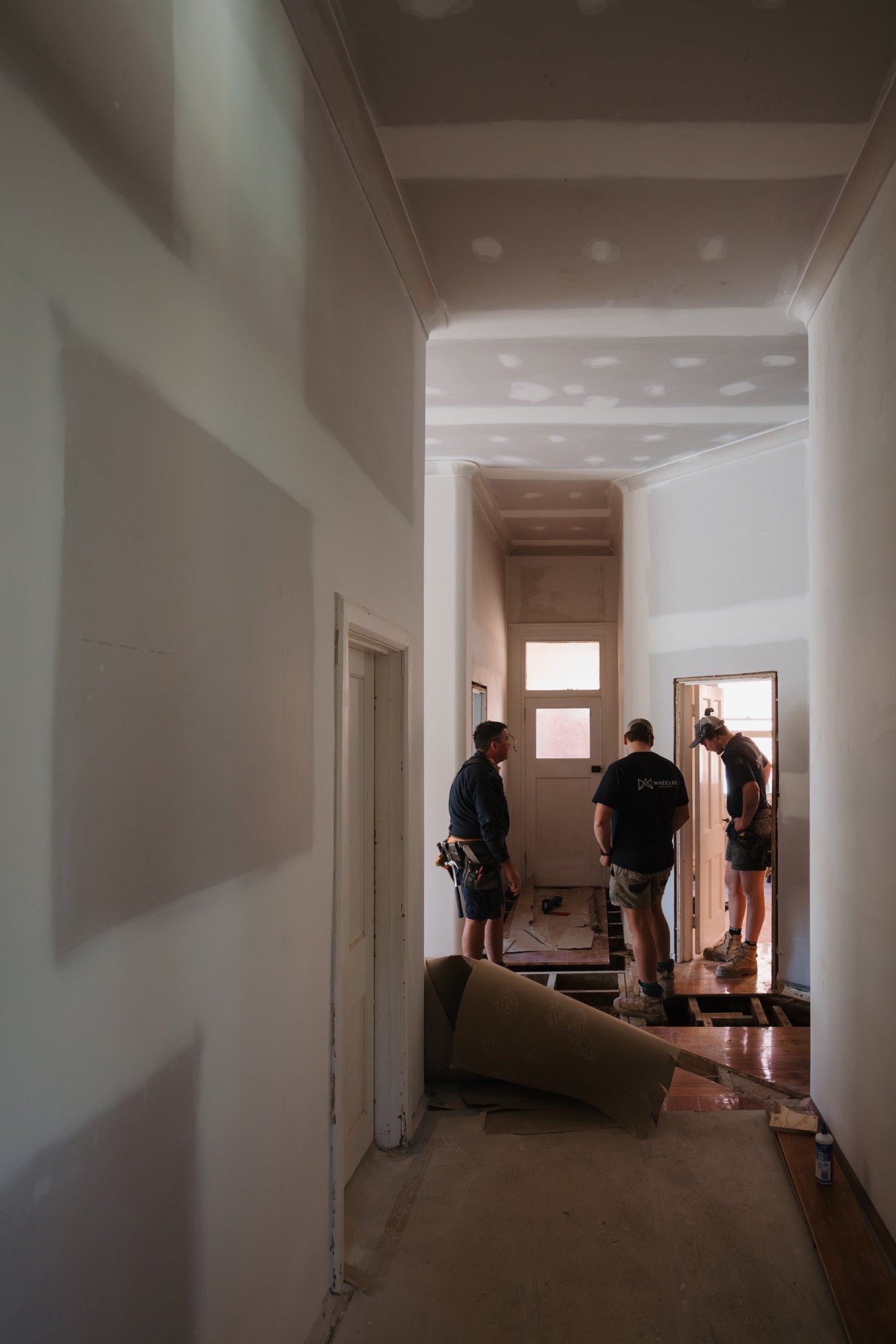 Construction workers inside a house under renovation, working near a doorway with a partially finished floor and ceiling with visible drywall seams.