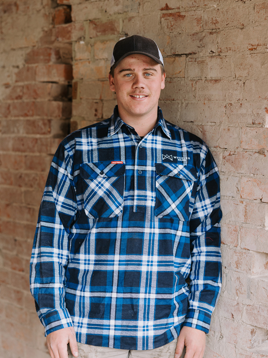 Young man wearing a black and white trucker cap, blue and black plaid shirt, standing against a brick wall.