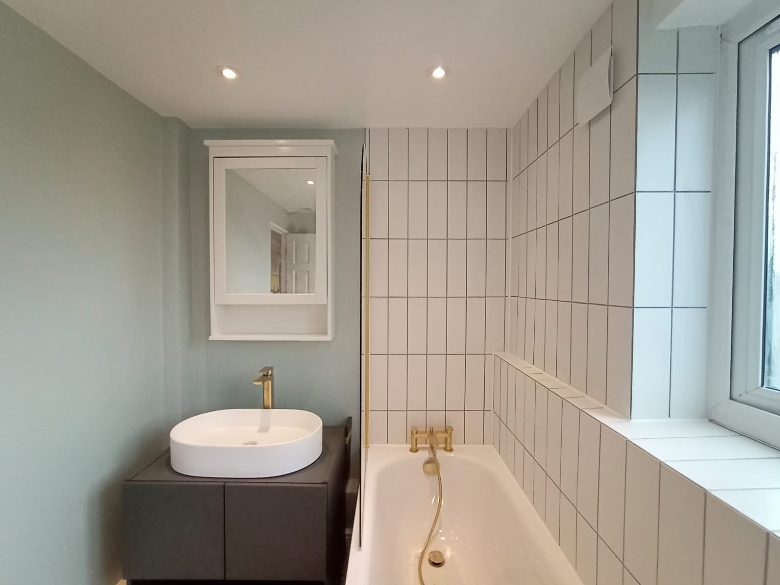A newly renovated bathroom with a white bathtub, a white sink with gold fixtures, a cabinet with a mirror, and a subway tiles around the window.