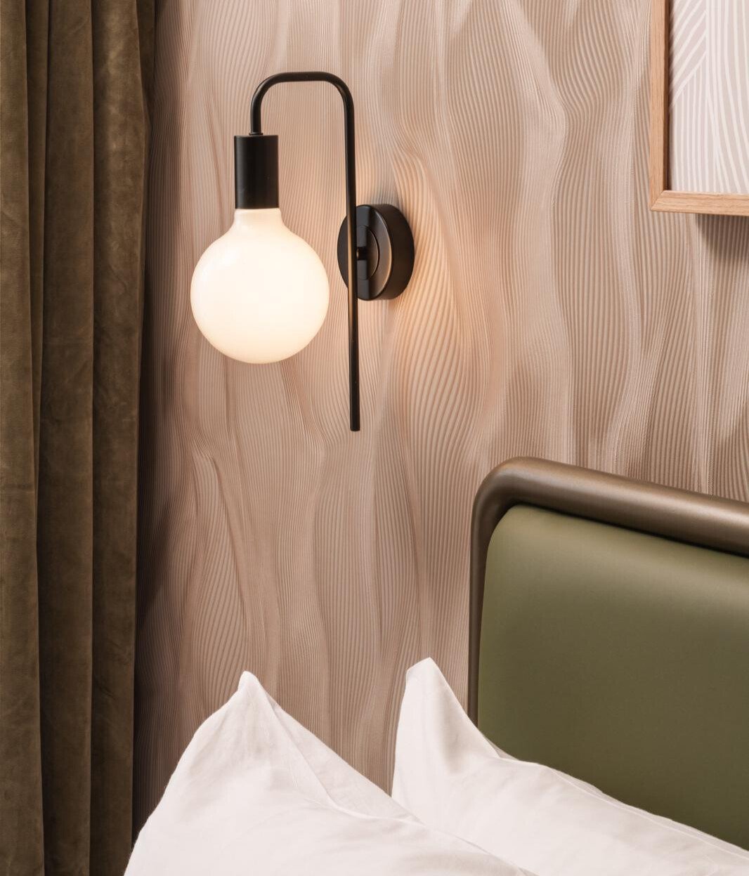 A newly-renovated and redesigned bedroom with wall-mounted modern light fixture with a black metal arm and a frosted white globe bulb, next to a bed with white bedding and a green upholstered headboard.