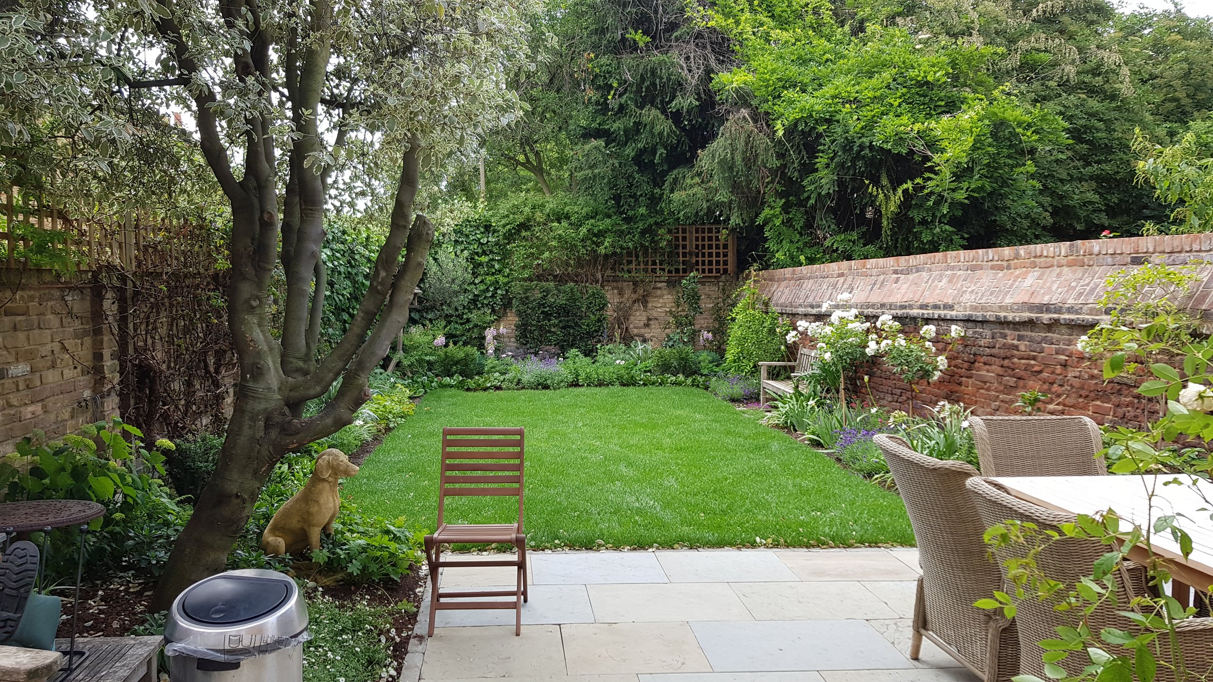 A back garden with a lawn, surrounded by trees, shrubs, and flowers. There is a brick wall on the right side that has been restored and re-enforced.