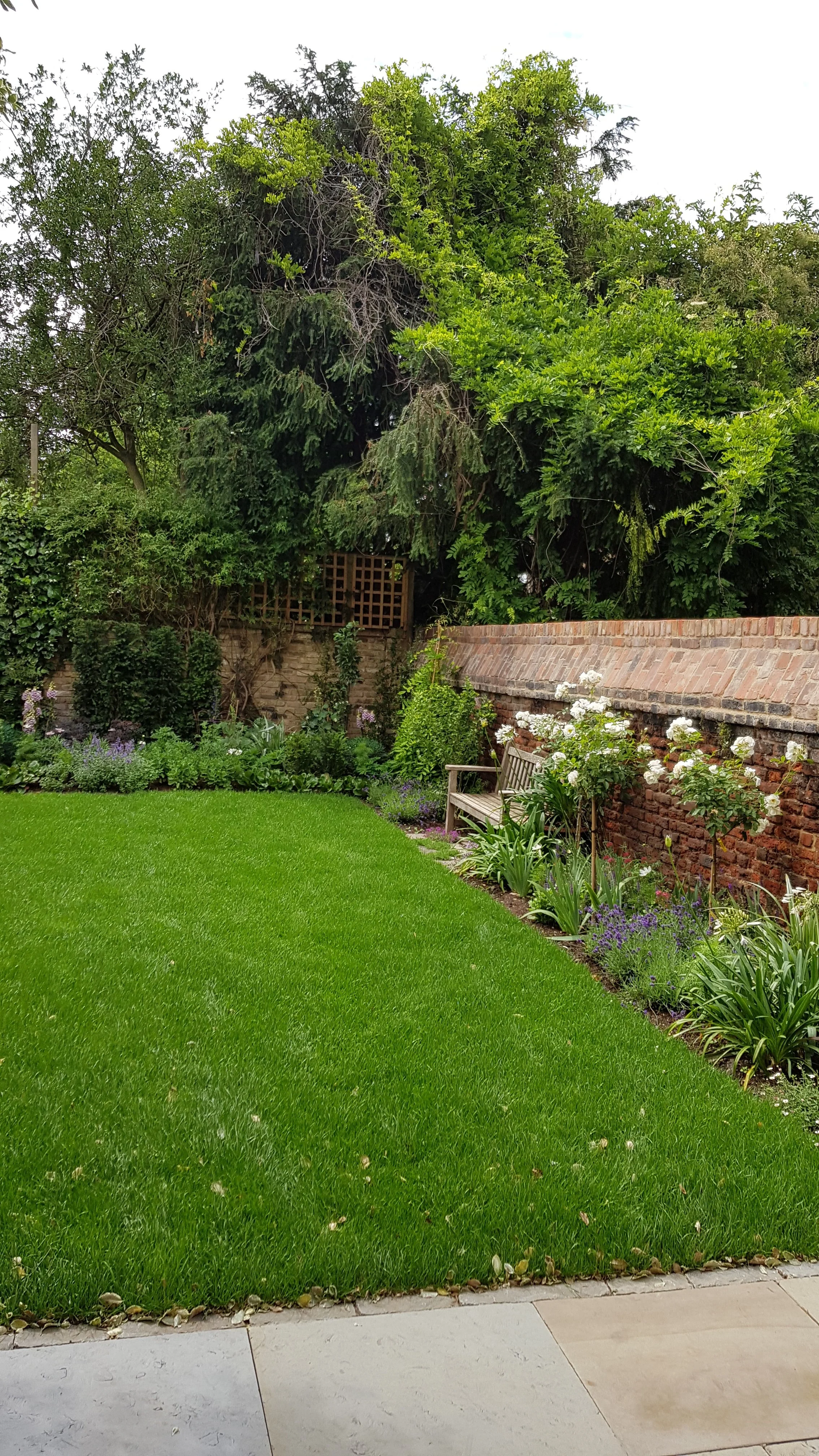 A lush green garden with a neatly trimmed lawn, a wooden bench along a brick wall, and various flowering plants and bushes, with trees in the background.