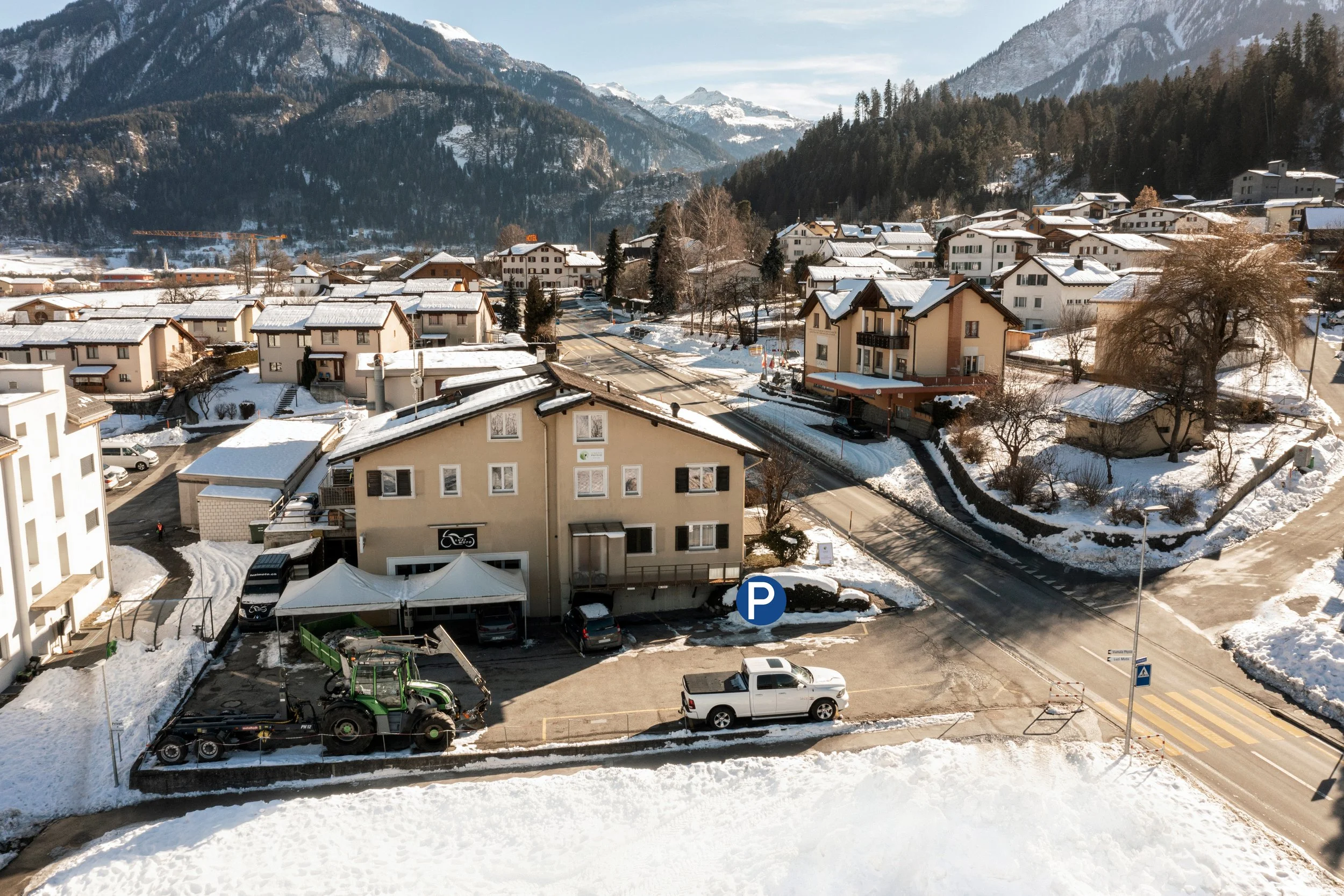 Schneebedeckte Häuser in Sumaprada mit verschneiten Bergen im Hintergrund, Autos und ein Parkplatz im Vordergrund.