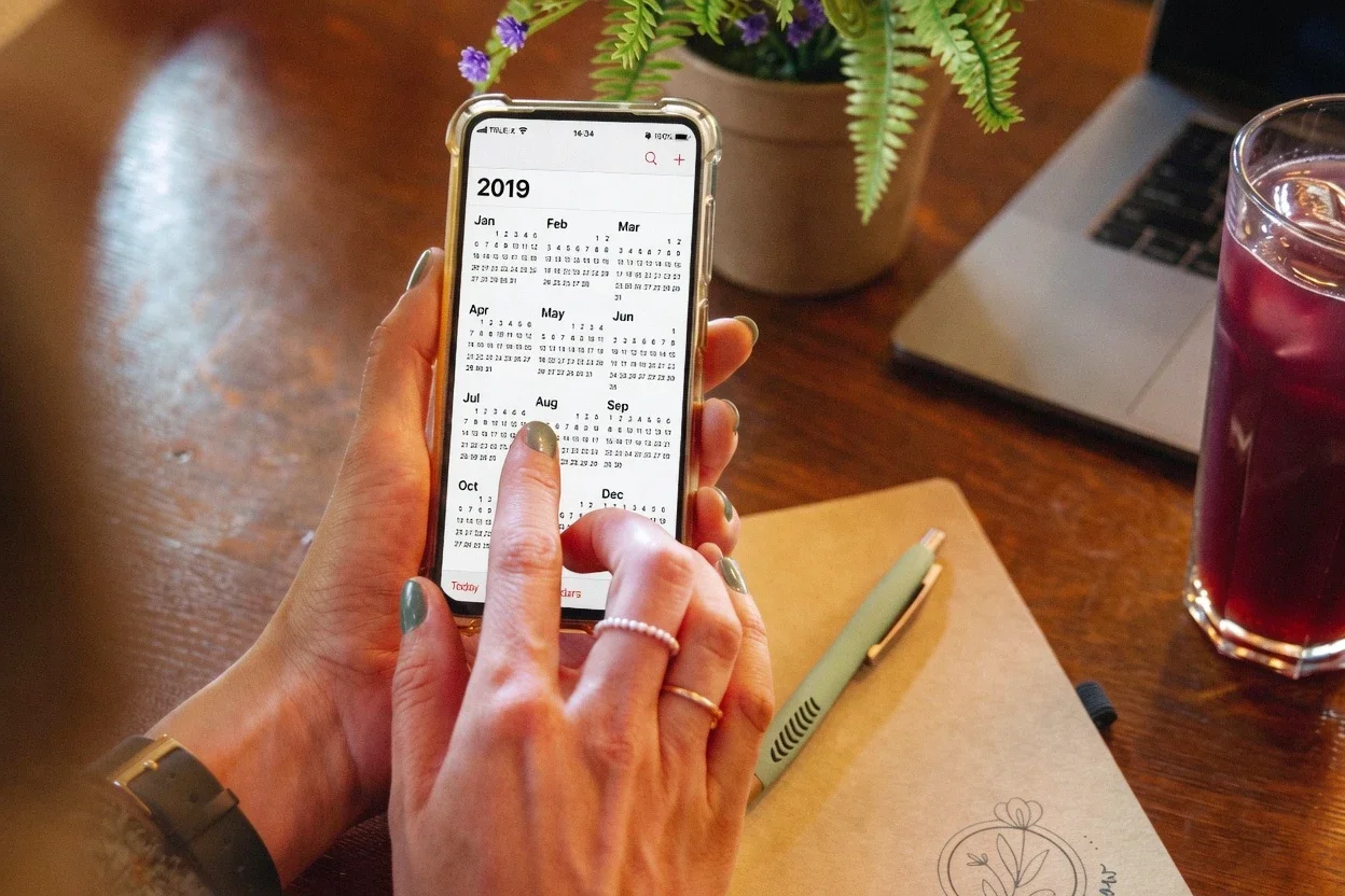 Person hält Smartphone mit einem Kalender . Auf dem Tisch liegen ein notizbuch, ein Stift und ein Glas mit einem dunklen Getränk. Im Hintergrund ist ein Laptop und eine Topfpflanze zu sehen. Jetzt Termin bei Mindful Growing für Achtsamkeitstrainig