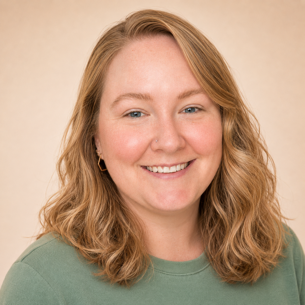 A woman with shoulder-length wavy red hair, blue eyes, and a nose piercing, smiling and wearing a green shirt and gold earrings.