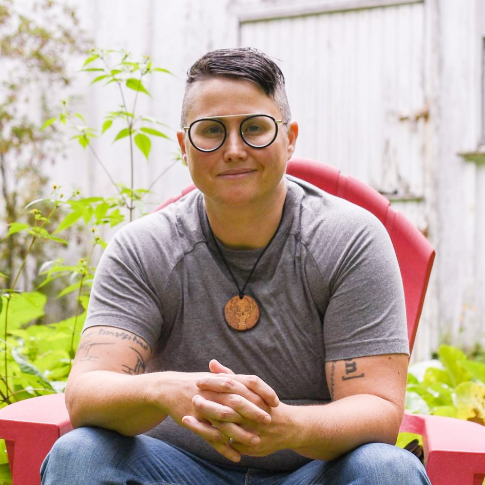 A person with short dark hair, glasses, and tattoos on their arms is sitting outside on a red chair with greenery and a white wooden fence in the background. They are wearing a gray t-shirt and a necklace with a wooden pendant.