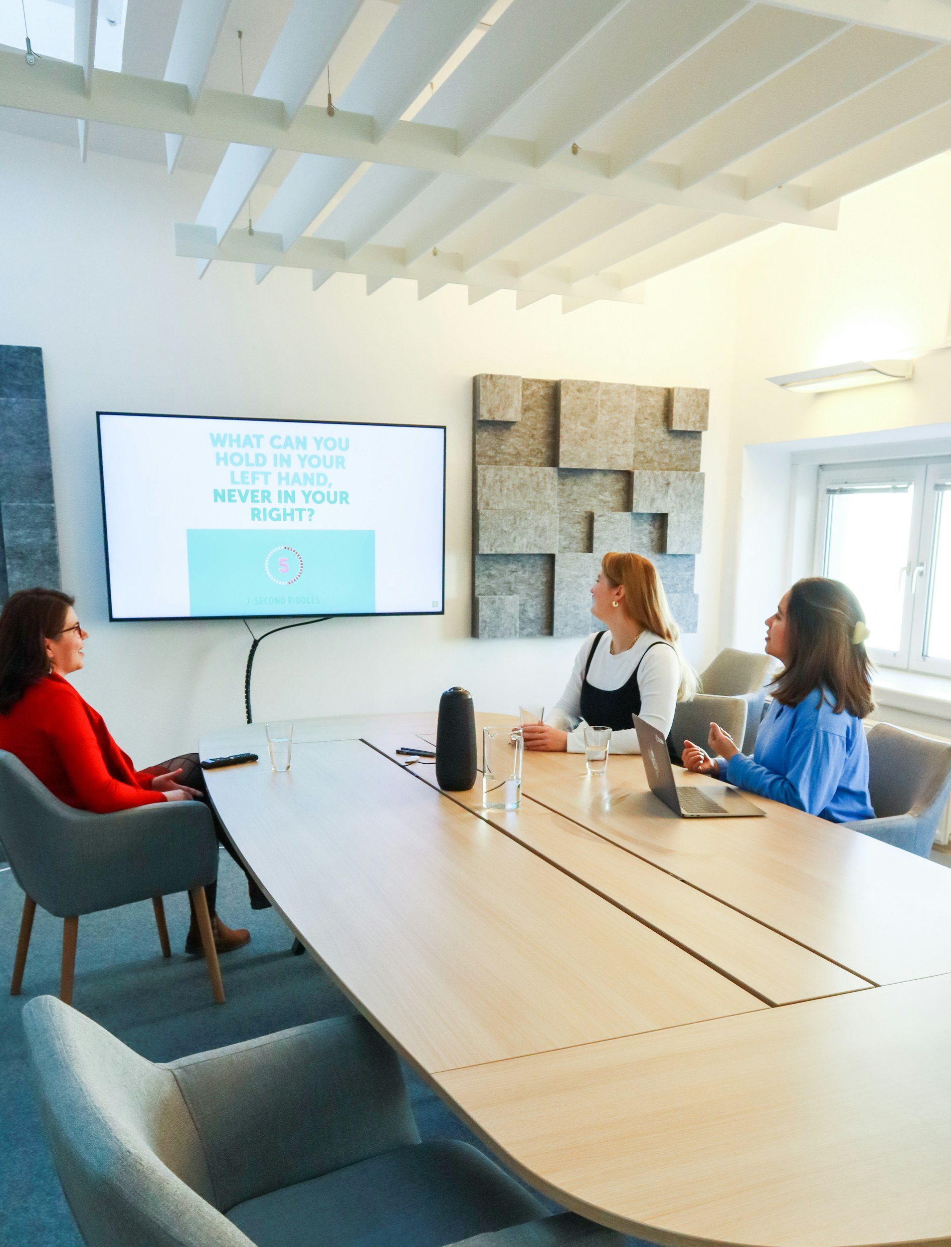 Three women sitting around a conference table in a meeting room, watching a presentation on a wall-mounted screen that shows a quiz question about holding something in the hands. One woman has a laptop, and there are glasses and a speaker on the table.