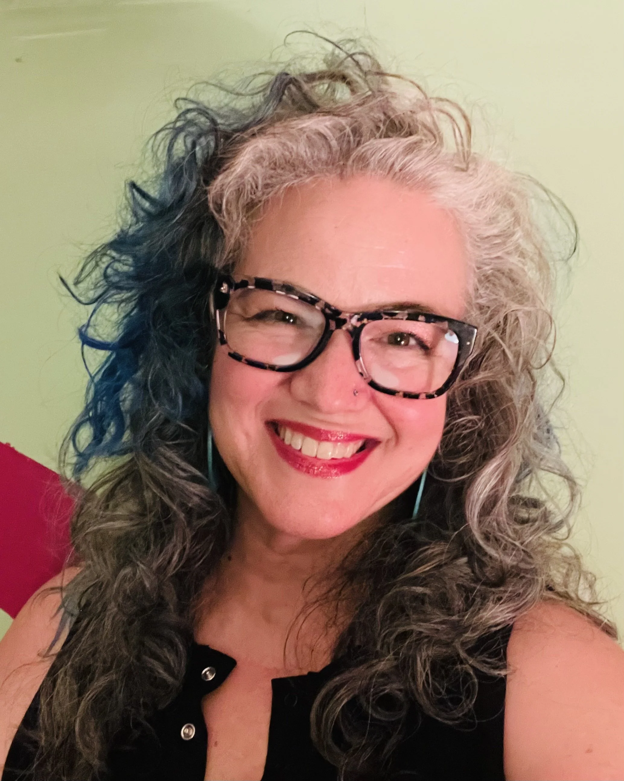 A woman with curly, two-toned hair, wearing glasses with a tortoiseshell pattern, smiling with red lipstick, and wearing a black top with metallic button details.
