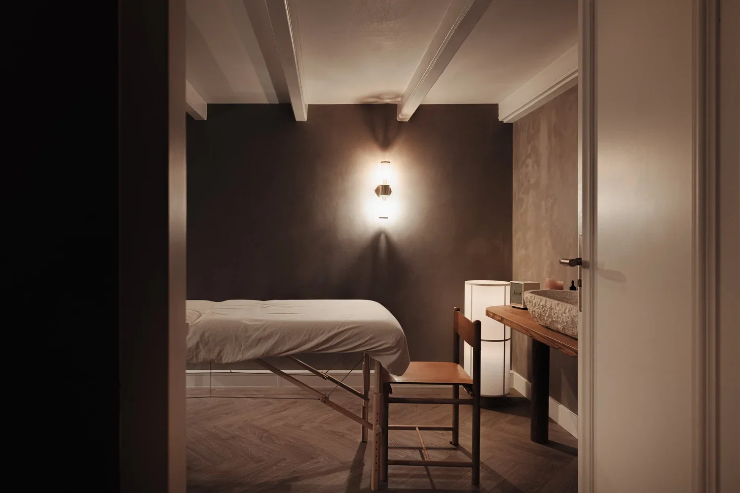 A minimalistic massage room with a massage table covered with white sheet, a wooden chair, a white bedside lamp, and a stone sink on a wooden countertop. The room has dark and neutral-colored walls, wood flooring, and soft warm lighting.