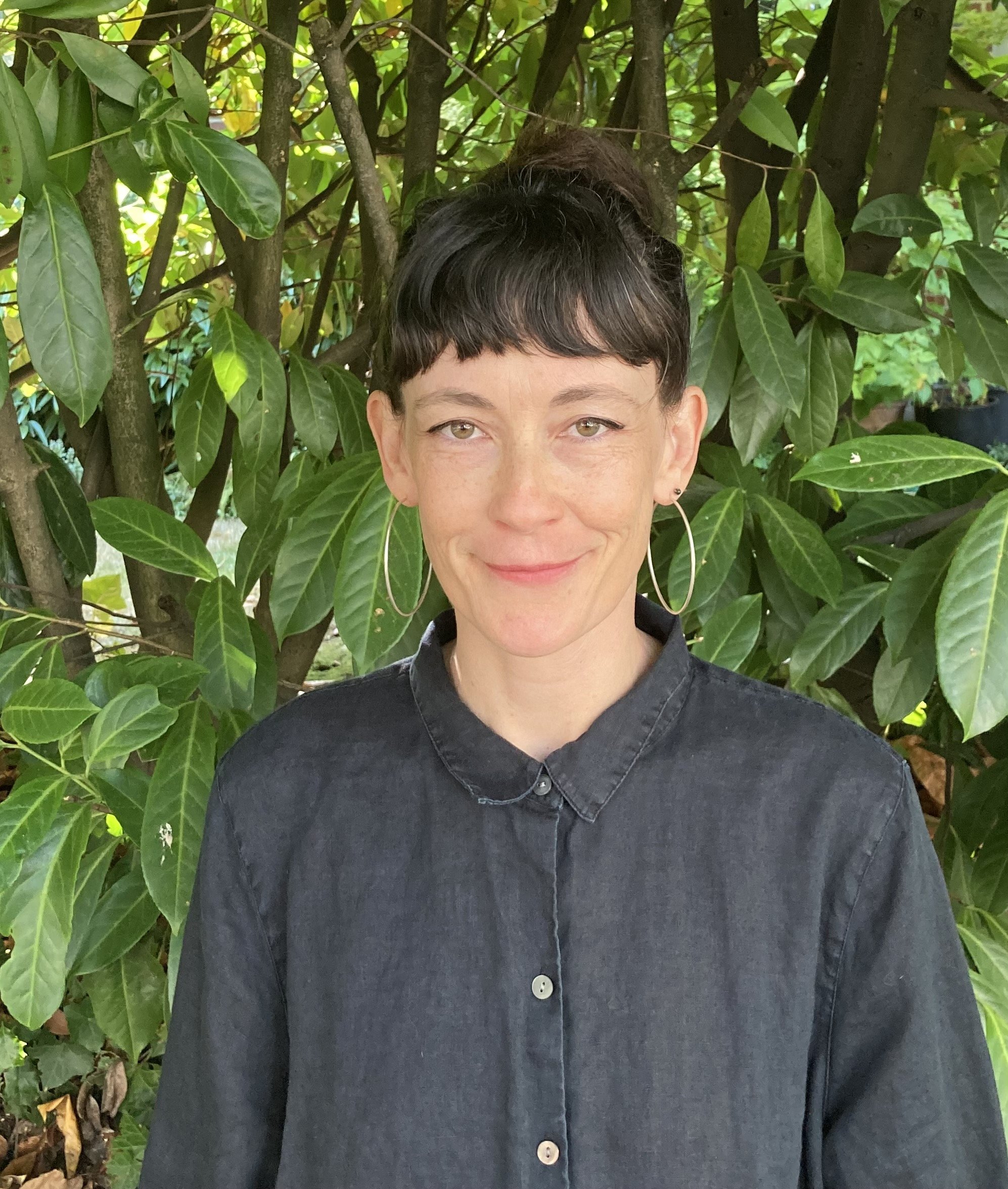 A woman with short dark hair and large hoop earrings standing outdoors in front of a leafy green bush, wearing a dark button-up shirt and smiling gently.