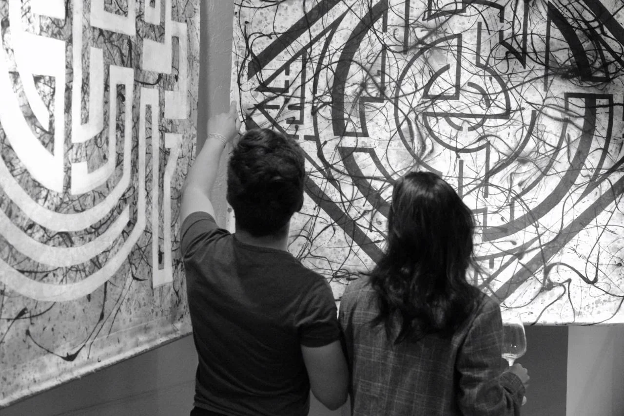 Two people are standing in front of abstract black and white artwork, one is pointing at the artwork while the other holds a wine glass.