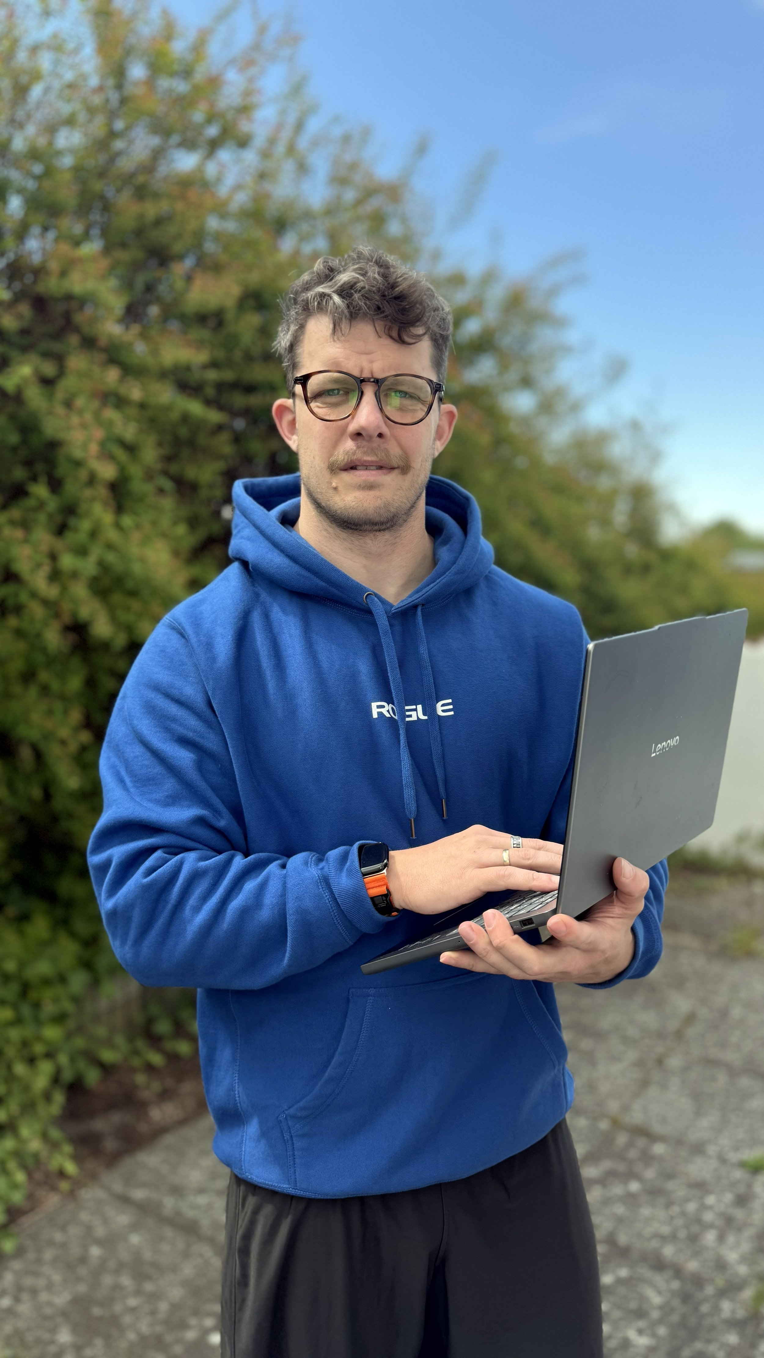 En mand, der står udenfor med en laptop i hånden, iført en blå hoodie og et ur på venstre hånd, omgivet af grønne buske mod en blå himmel.