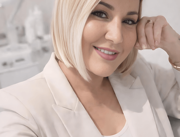 A woman with blonde hair smiling, wearing a white blazer, in an indoor setting.