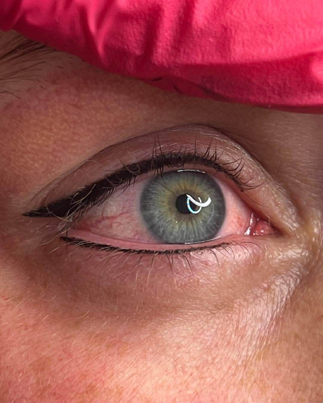 Close-up of a blue-green eye with makeup and reflections in the pupil, partially covered by pink fabric.