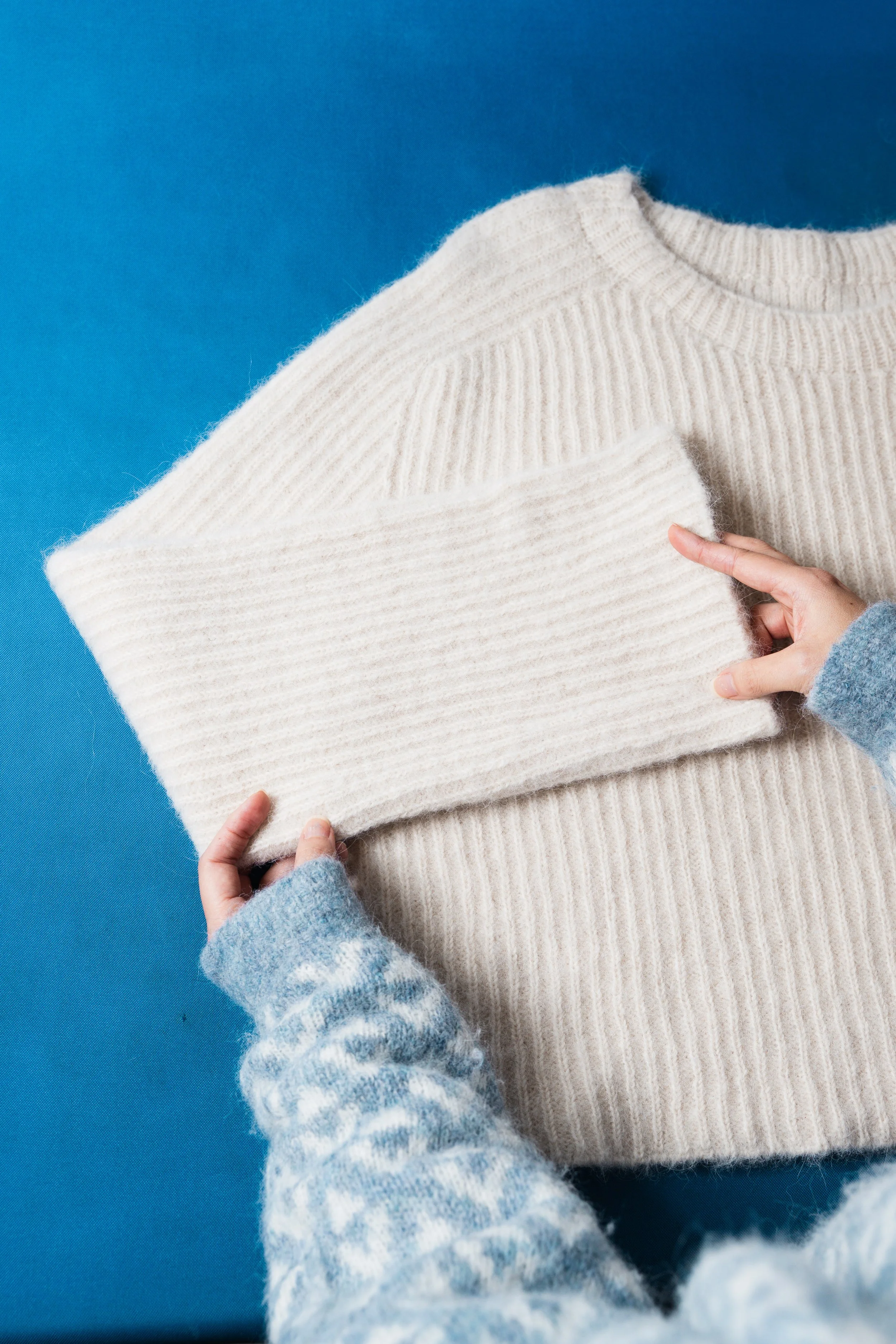 Produktfotografering av kvit strikkegenser mot blå bakgrunn, med fokus på tekstur og detaljar – visuelt innhald for bedrifter.