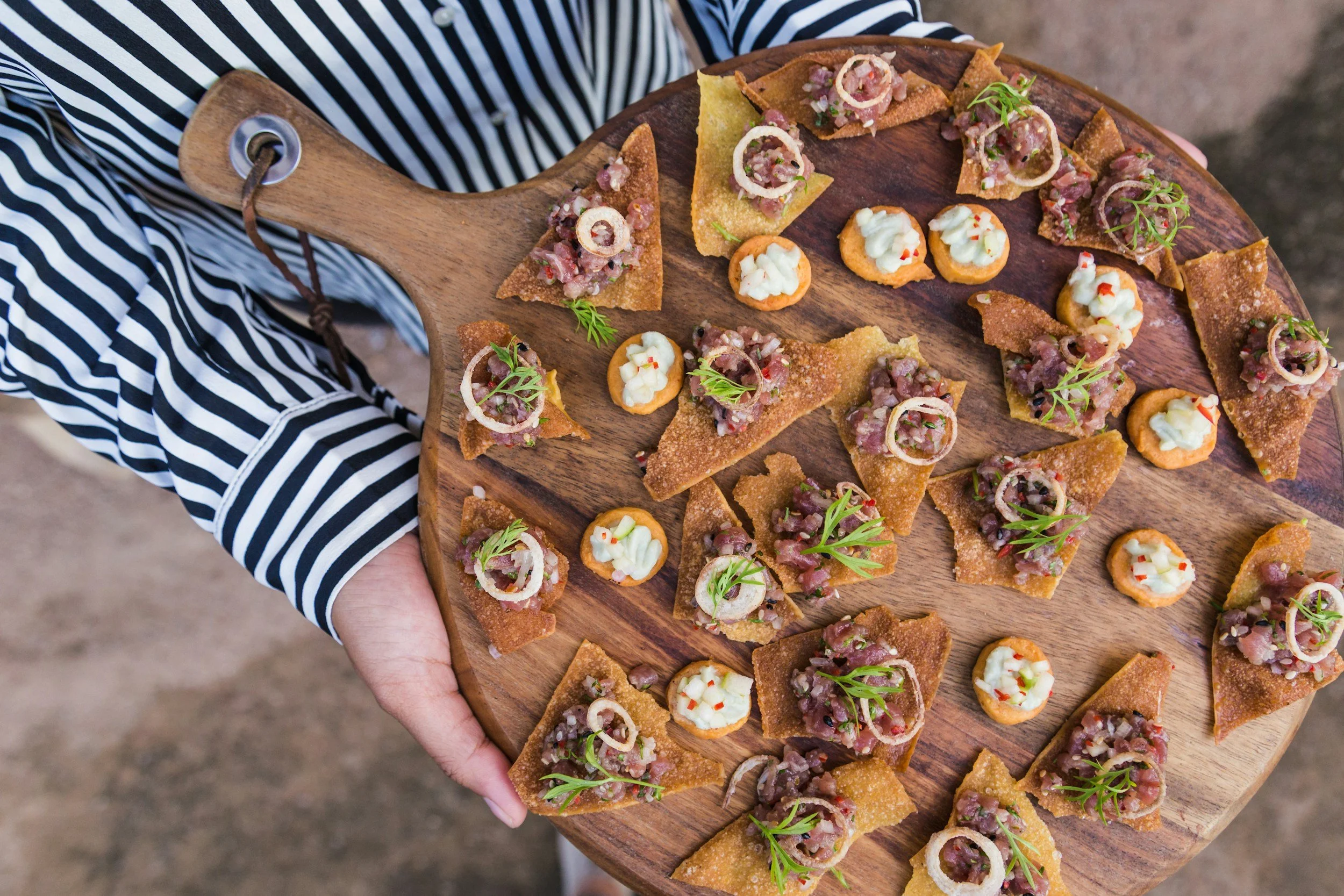 A person holding a wooden serving board with various appetizers, including small toasts with minced meat and onion slices, and others with a creamy topping, some garnished with herbs.