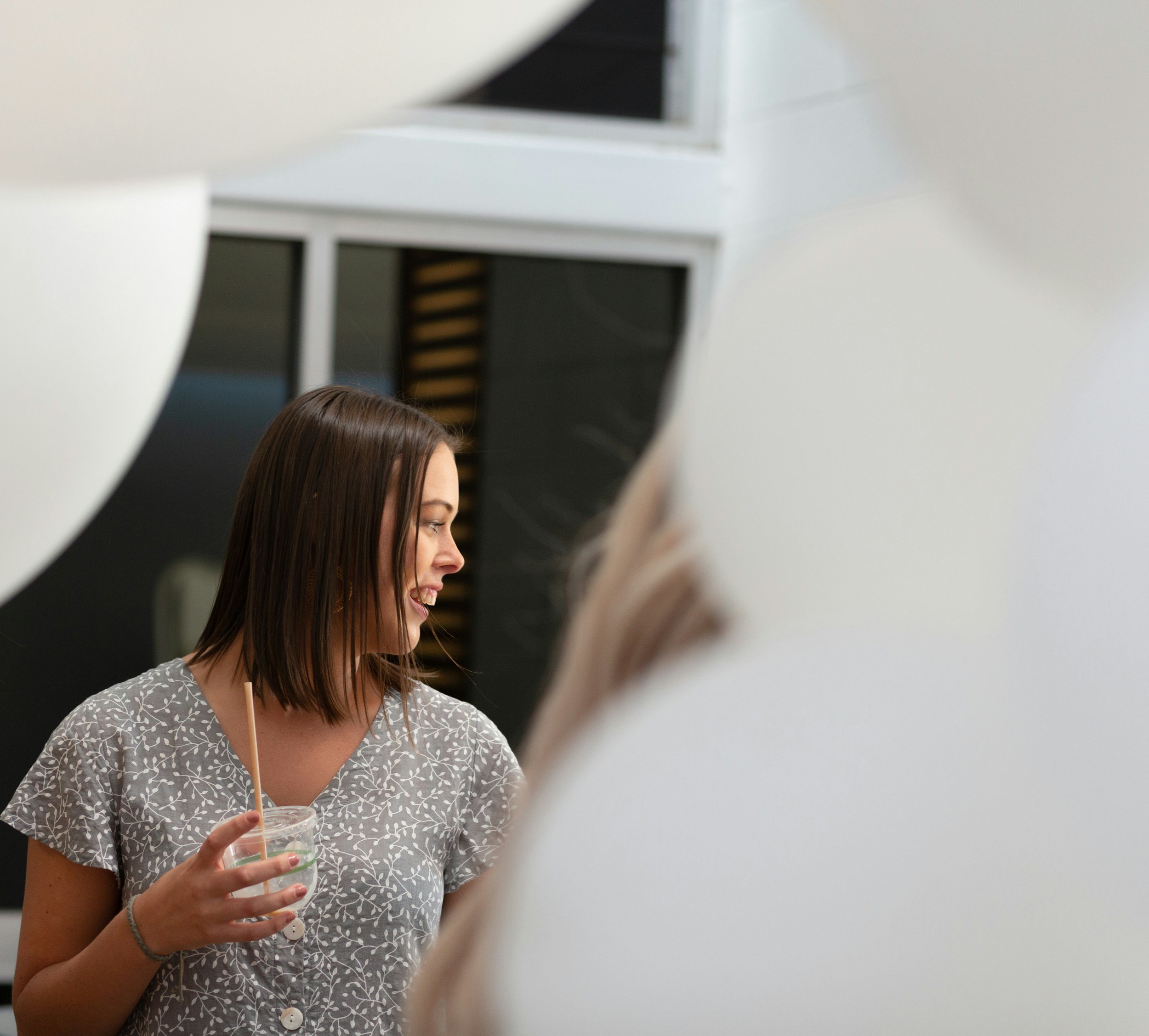 A woman with long brown hair holding a plastic cup with a straw, smiling and looking to the right, wearing a gray shirt with white geometric pattern, standing indoors.