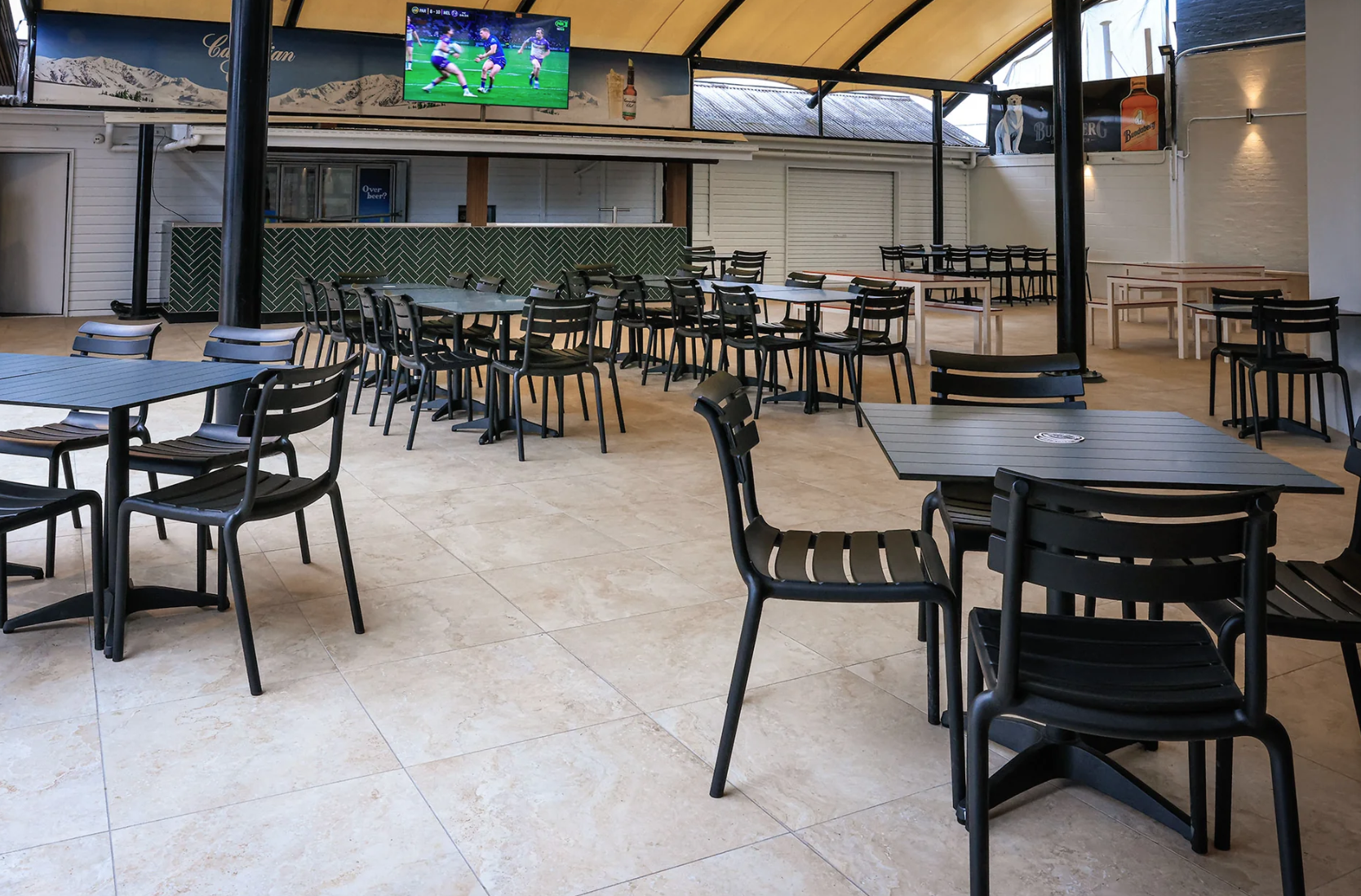 Empty indoor restaurant or cafe with black tables and chairs, TV screen showing a rugby game, and a green tiled counter.
