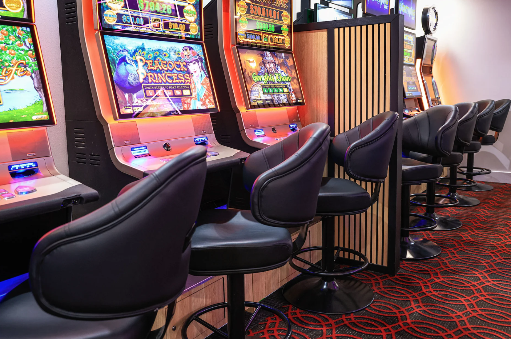 Row of electronic slot machines with black leather chairs and a patterned red carpet.