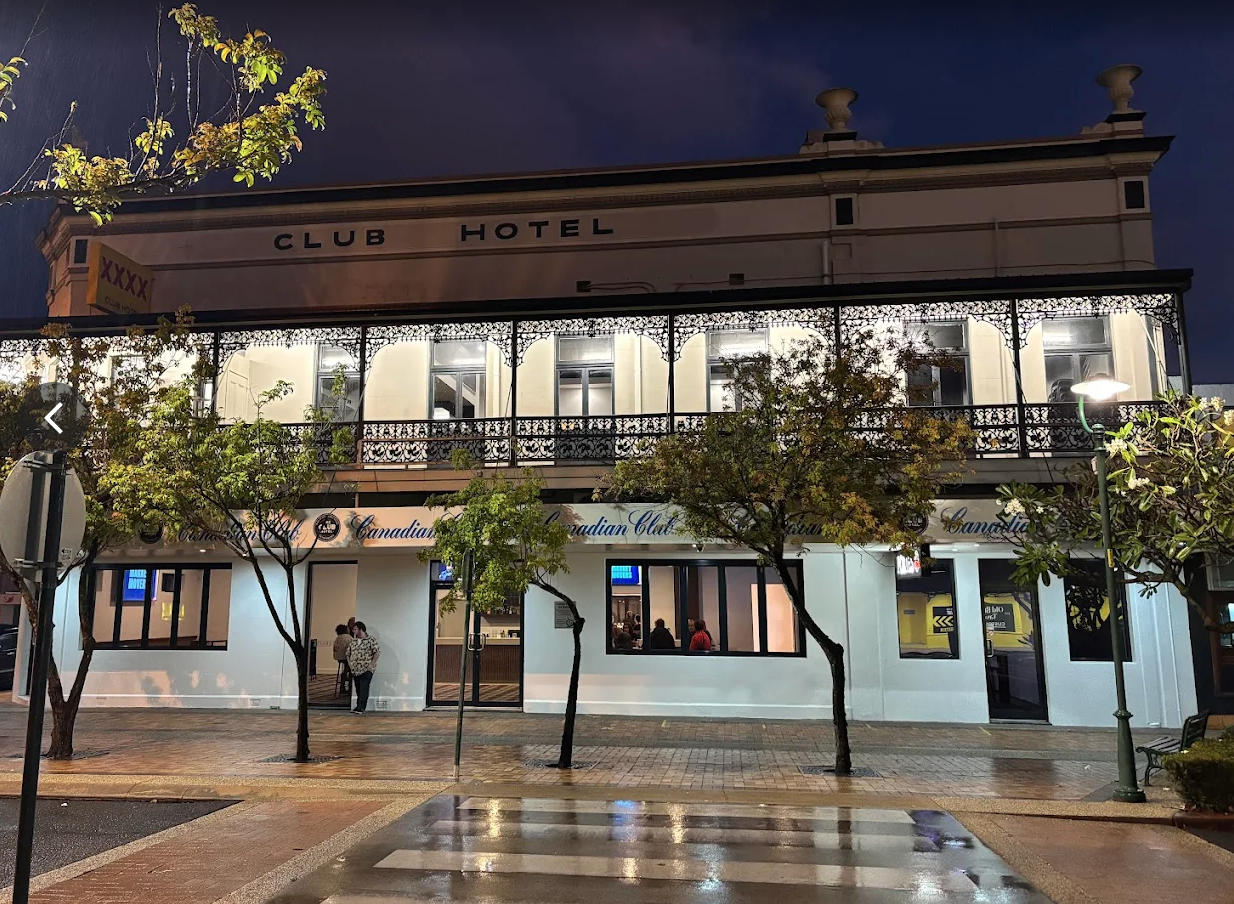 A two-story building at night with the sign 'Club Hotel' on the upper level. The lower level has a Canadian Club sign and a few people standing near the entrance. The street in front has trees, a crosswalk, and streetlights, with the sidewalk wet from rain.