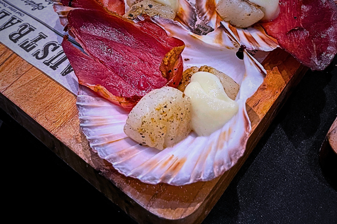 Assorted seafood, including scallops and slices of cured meat, served on a shell-shaped plate on a wooden board