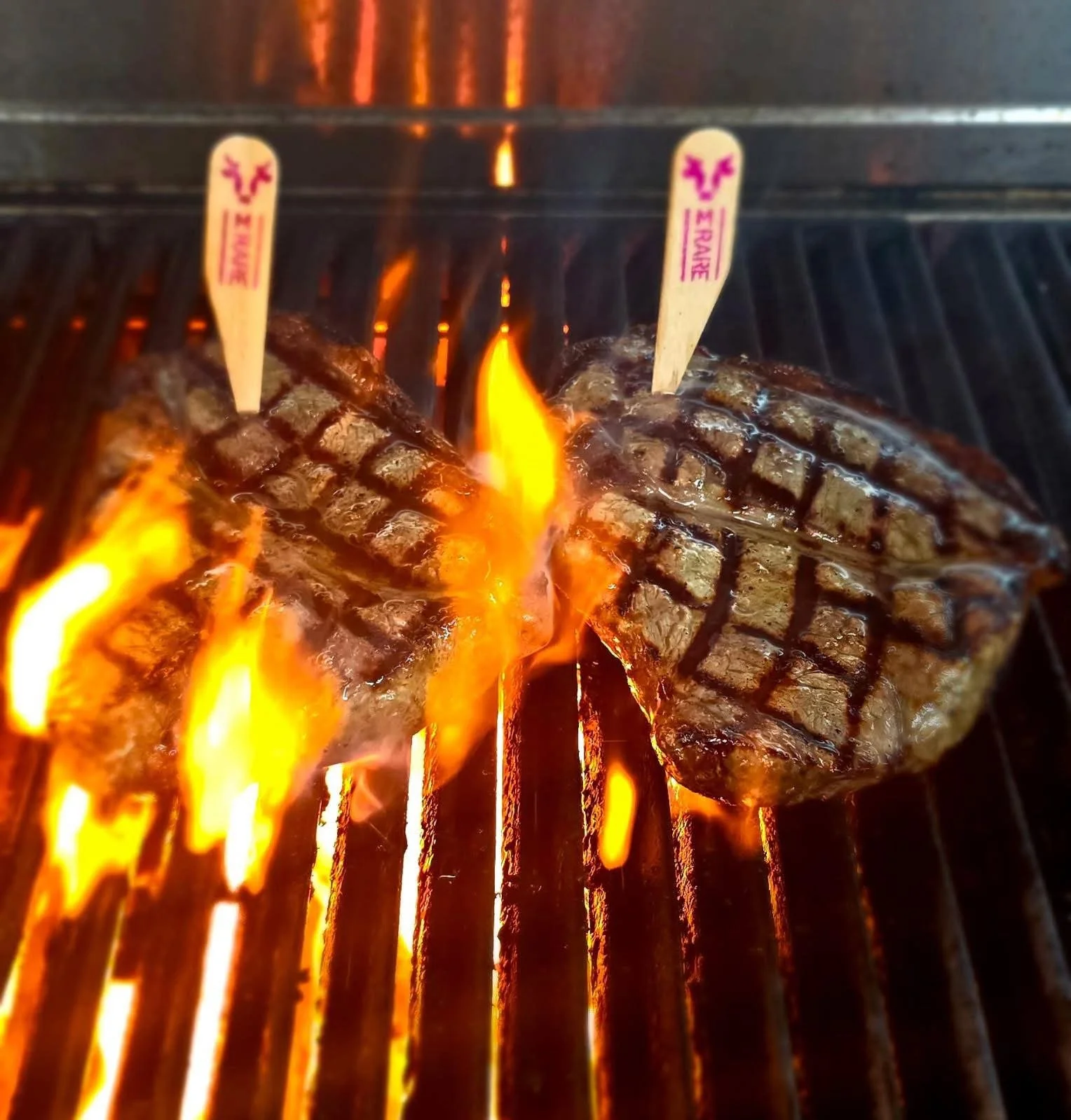 A piece of steak being grilled over open flames on a barbecue grill, with two small wooden skewers labeled 'E RAINFO' and 'E RAINB' inserted into it.