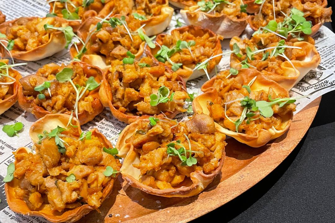 Stuffed pastry cups filled with seasoned diced vegetables and meat, garnished with small green microgreens on a wooden serving tray lined with newspaper.
