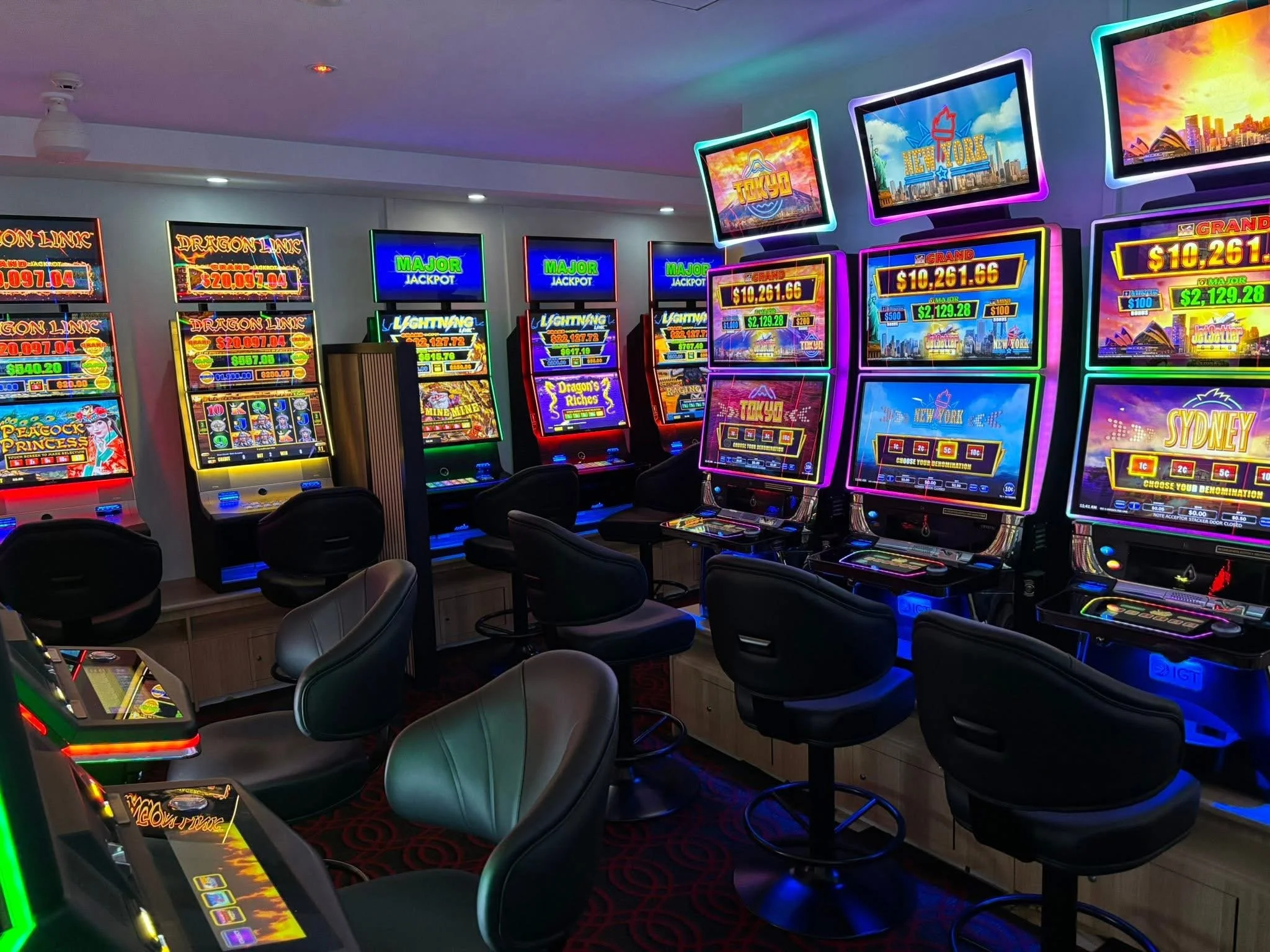 A room filled with colorful slot machines with bright lights, some showing large jackpots and game themes, accompanied by black chairs for players.