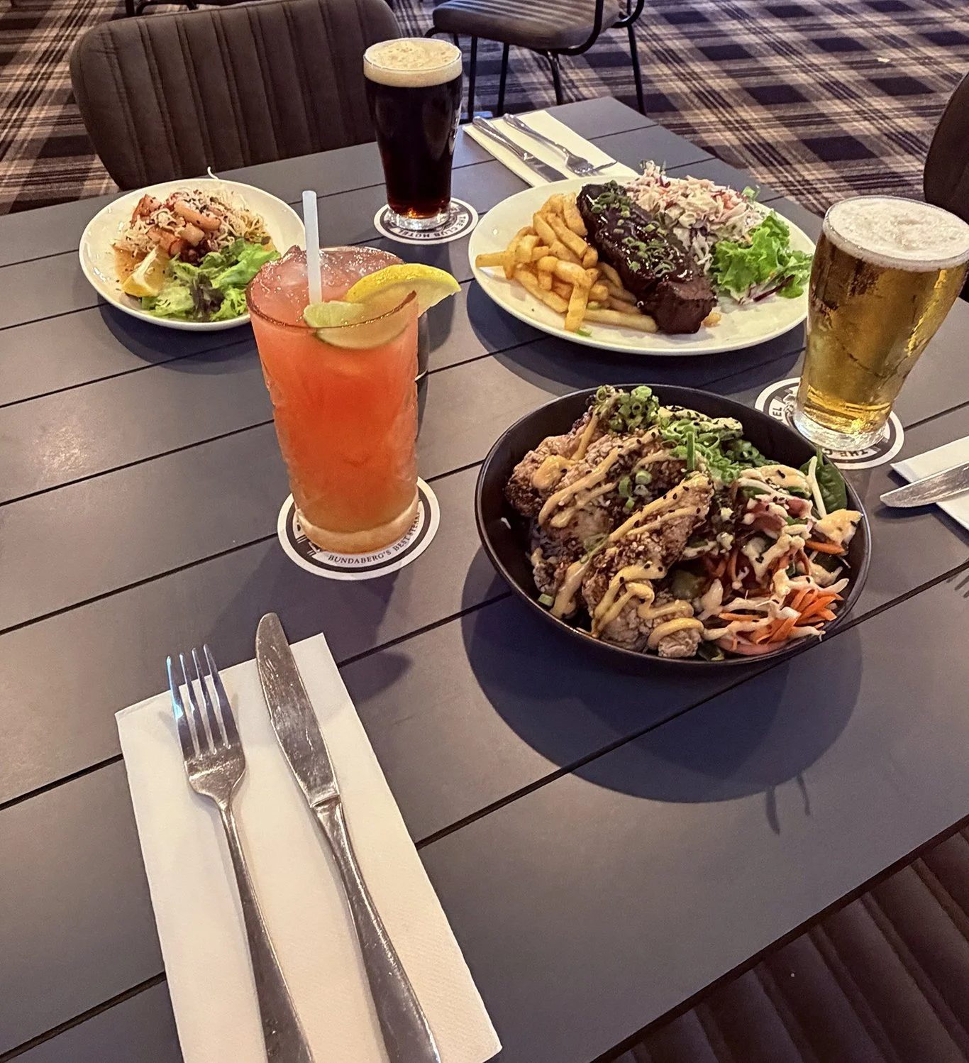 A table with a variety of foods and drinks including a salad, a barbecue plate with fries and coleslaw, a bowl of fried chicken topped with green onions, a pink cocktail with lemon slices, a pint of dark beer, and a lager.