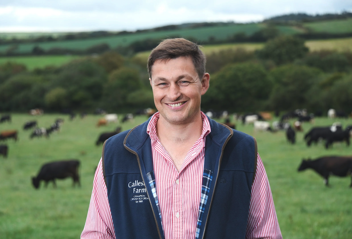 Smiling man standing in a green field with grazing cows and rolling hills in the background.