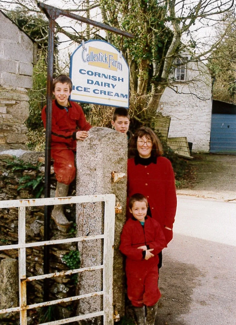 A woman and two children standing outside Callestick Farm, marked as a place that sells Cornish dairy ice cream. The children are wearing red clothes, and the woman is smiling. They are near a stone pillar and a white metal gate, with trees and buildings in the background.