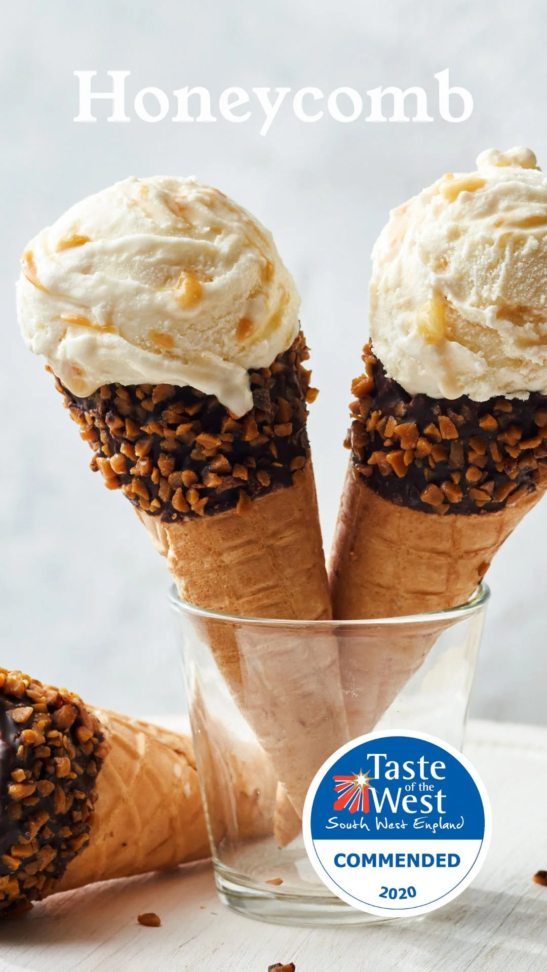 Two ice cream cones with vanilla ice cream, chocolate coating with chopped nuts, in a glass holder, with a third cone on a white surface and a "Taste of the West" award sticker.