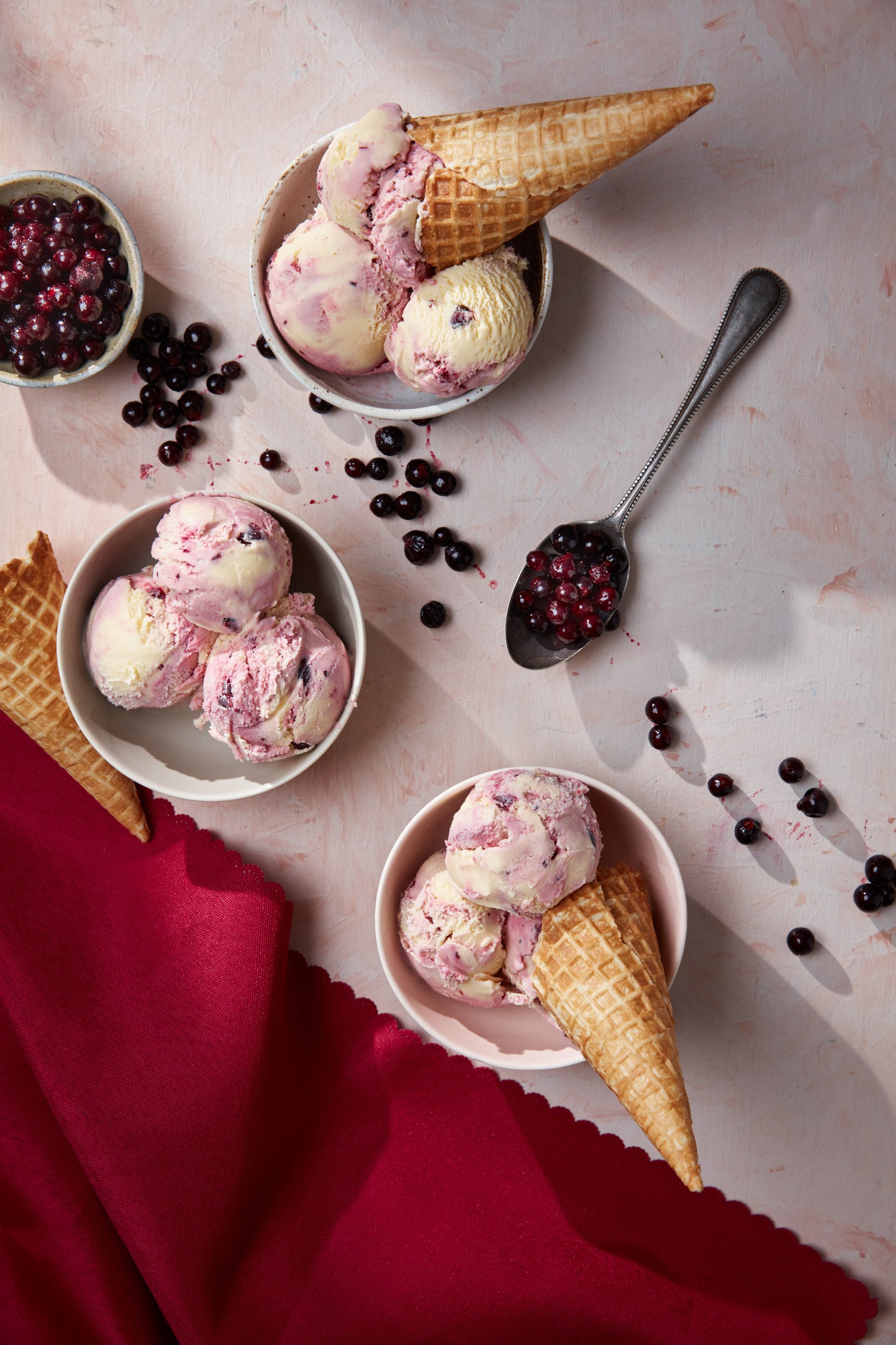 Three bowls of ice cream with pink and white swirls and waffle cones, scattered blueberries, and a spoon with blueberries on a light pink surface, with a red cloth in the bottom left corner.