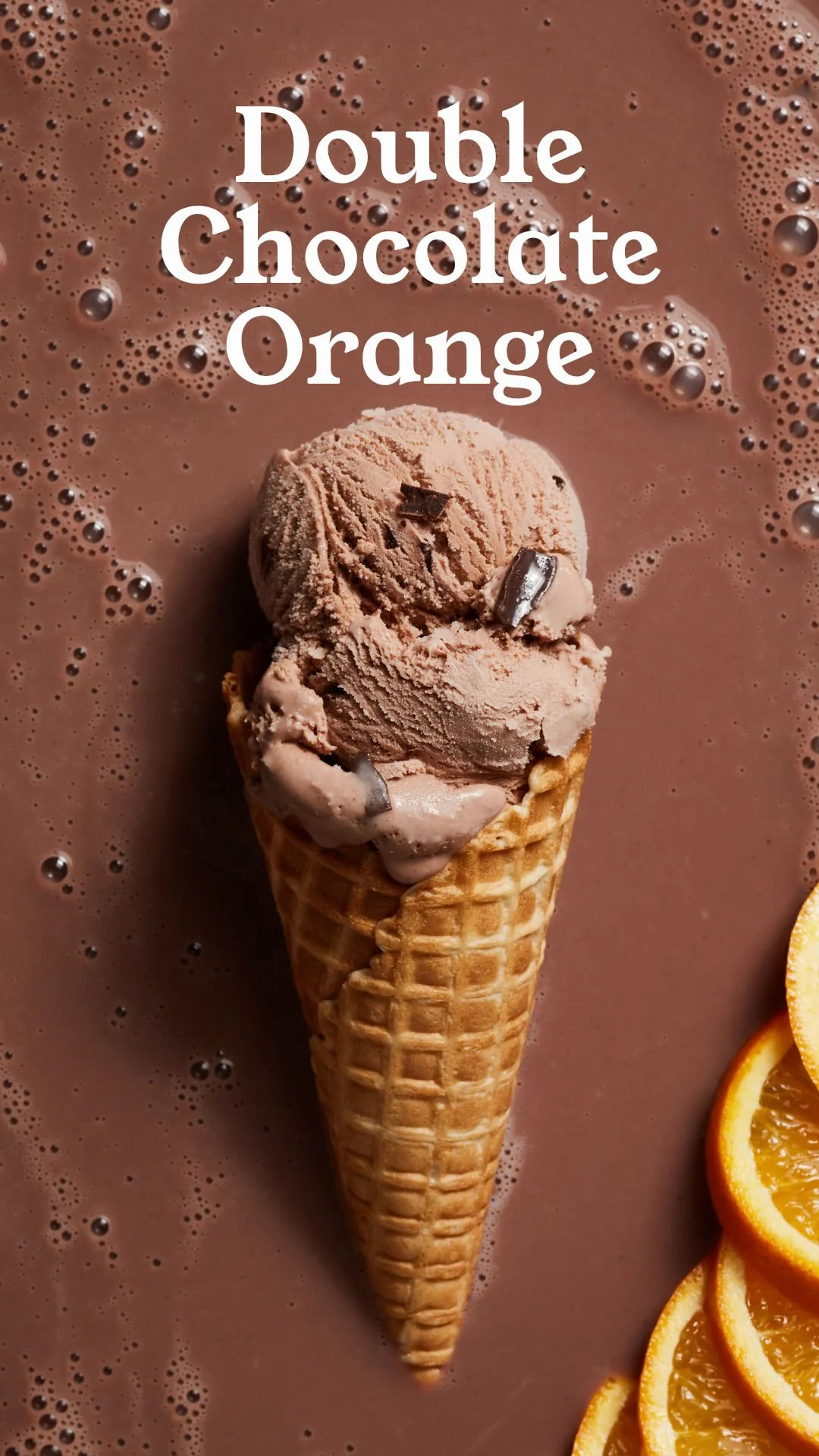 A scoop of chocolate ice cream with orange zest and chocolate shavings in a waffle cone sitting on a chocolate surface, with orange slices on the side.
