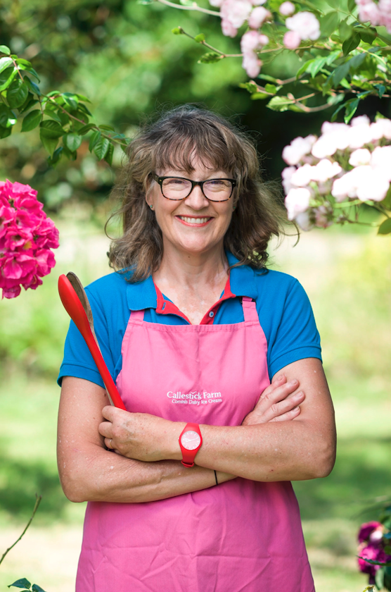 A woman smiling outdoors among blooming pink and white flowers, wearing glasses, a pink apron, and a blue shirt, holding a red gardening tool.