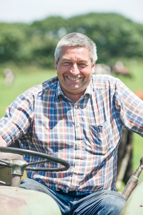 Smiling man sitting on a tractor outdoors in a field with green trees in the background.