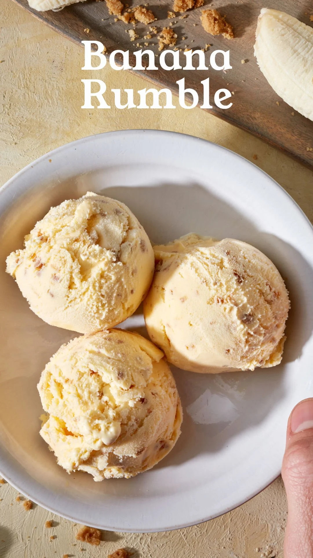 Three scoops of banana ice cream in a white bowl with a banana and cookie crumbs in the background.