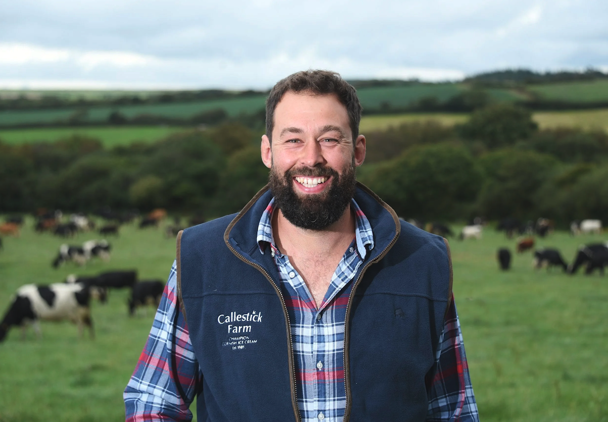 Smiling man with a beard standing in a field with cows and rolling green hills in the background.
