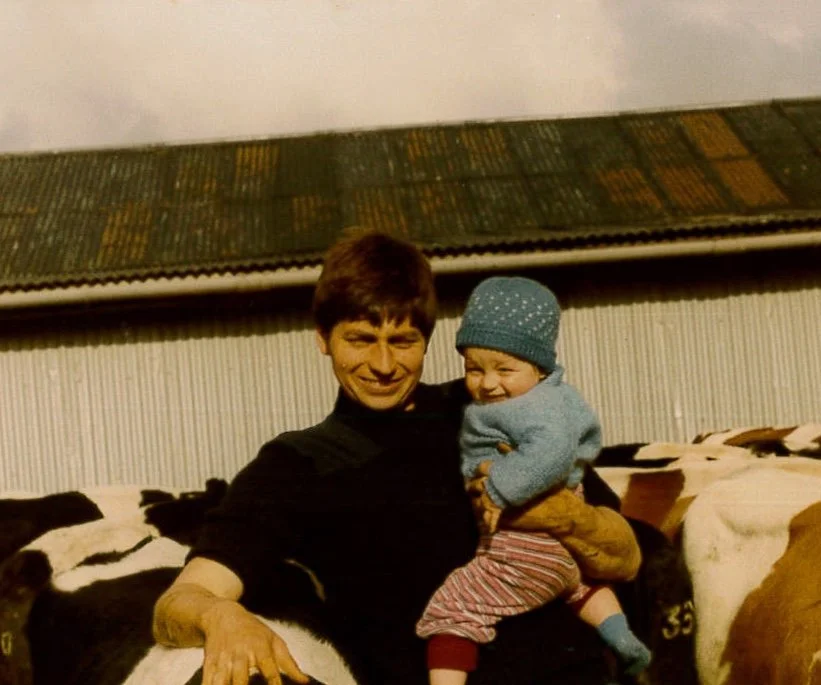 A woman holding a laughing child on her lap, with cows in the background and a building with a corrugated metal roof behind them.